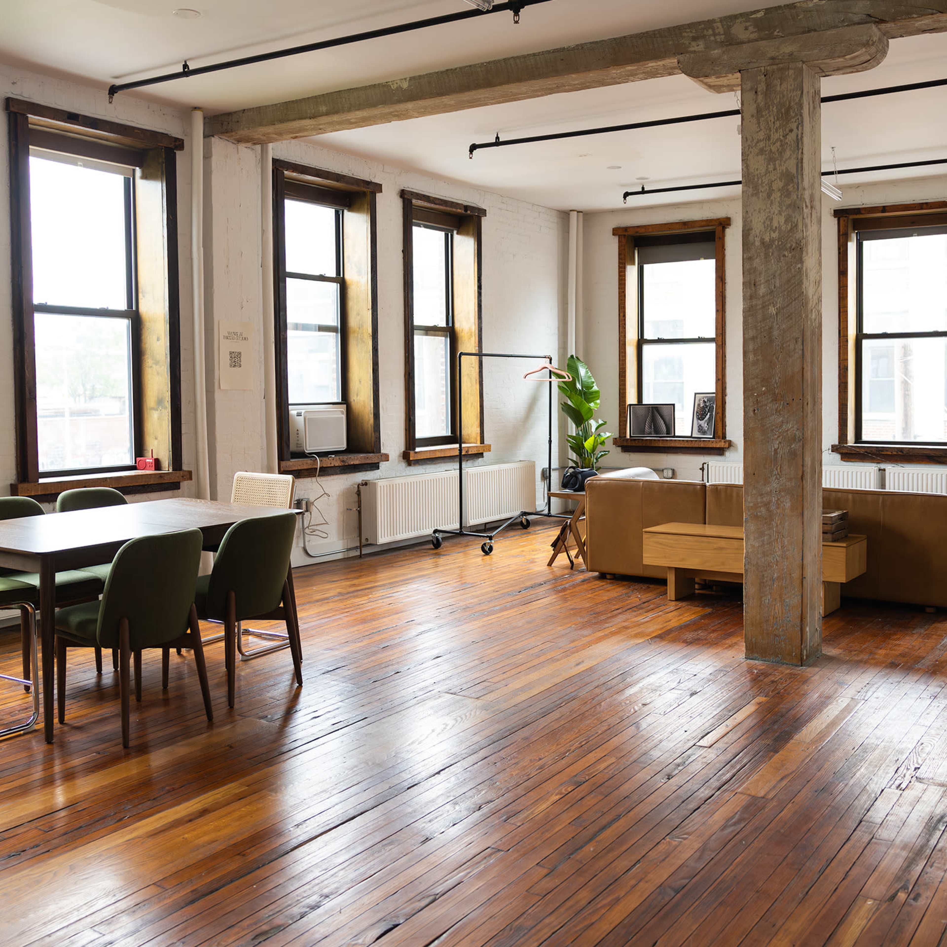 A spacious, empty room with large windows, wooden floors, a dining table with chairs, and a living area with a sofa and a plant.