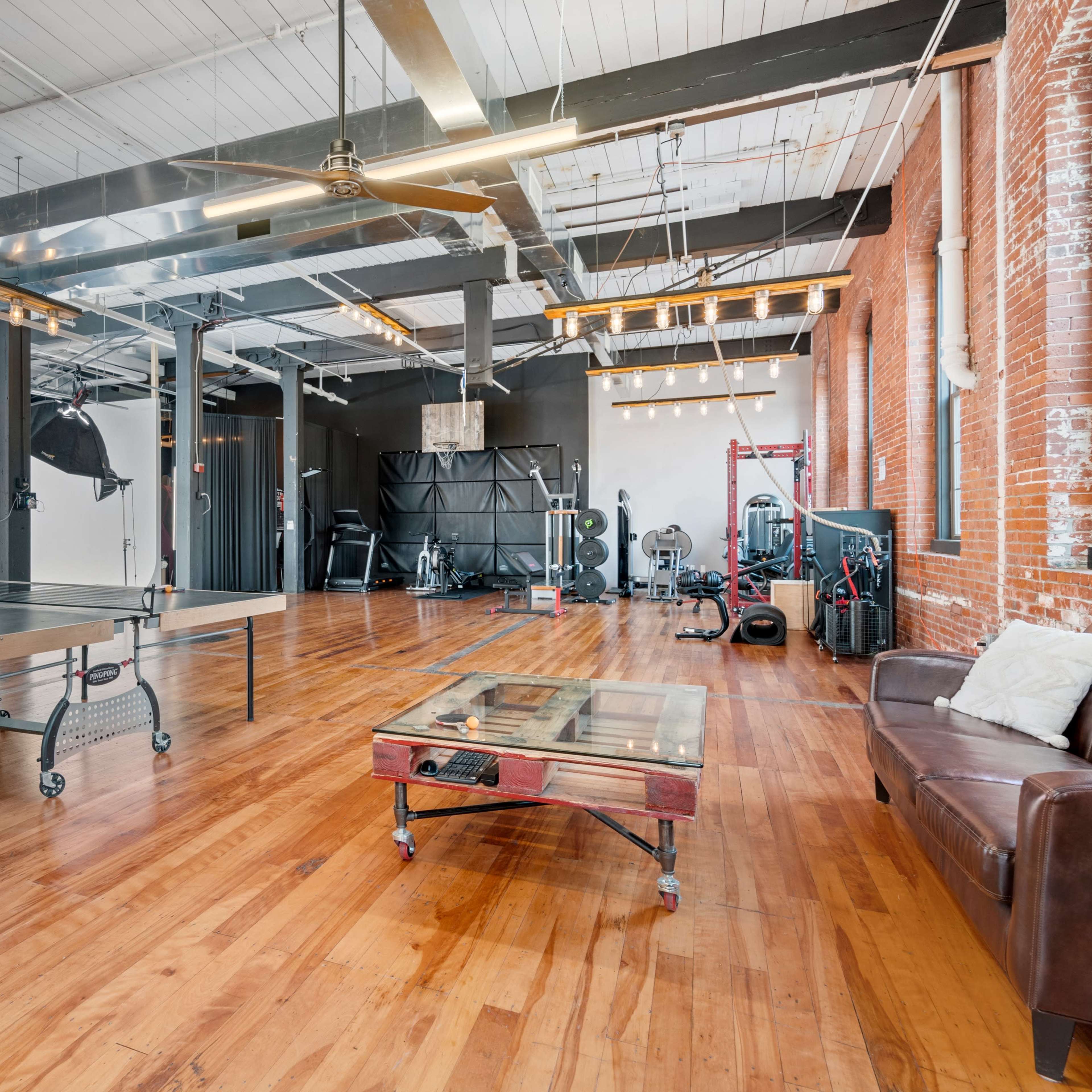A spacious industrial-style studio featuring polished wooden floors, exposed brick walls, and various workout equipment arranged in a well-lit area.
