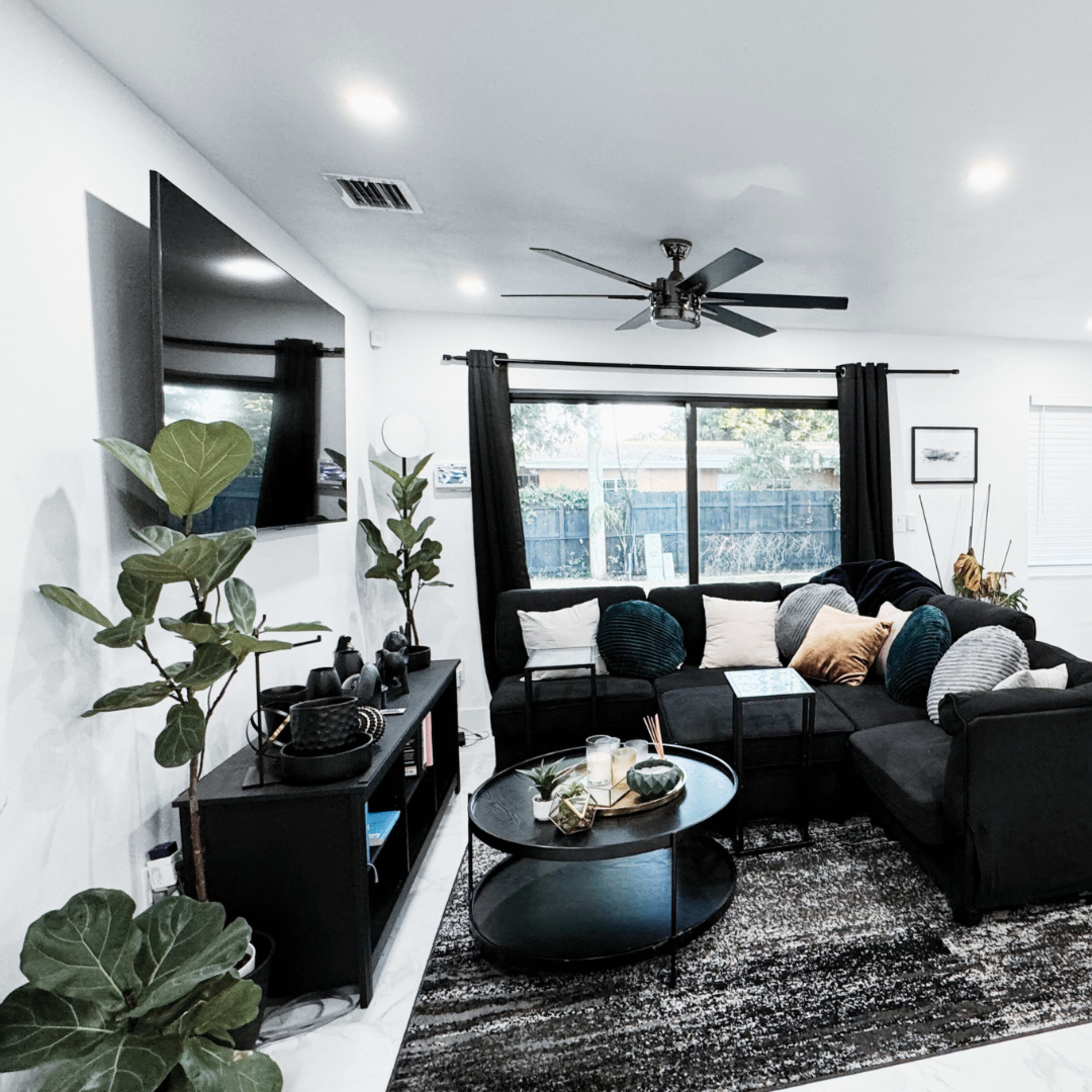 The living room features a black sectional sofa, a round coffee table, and a dining area with a large painting on the wall.