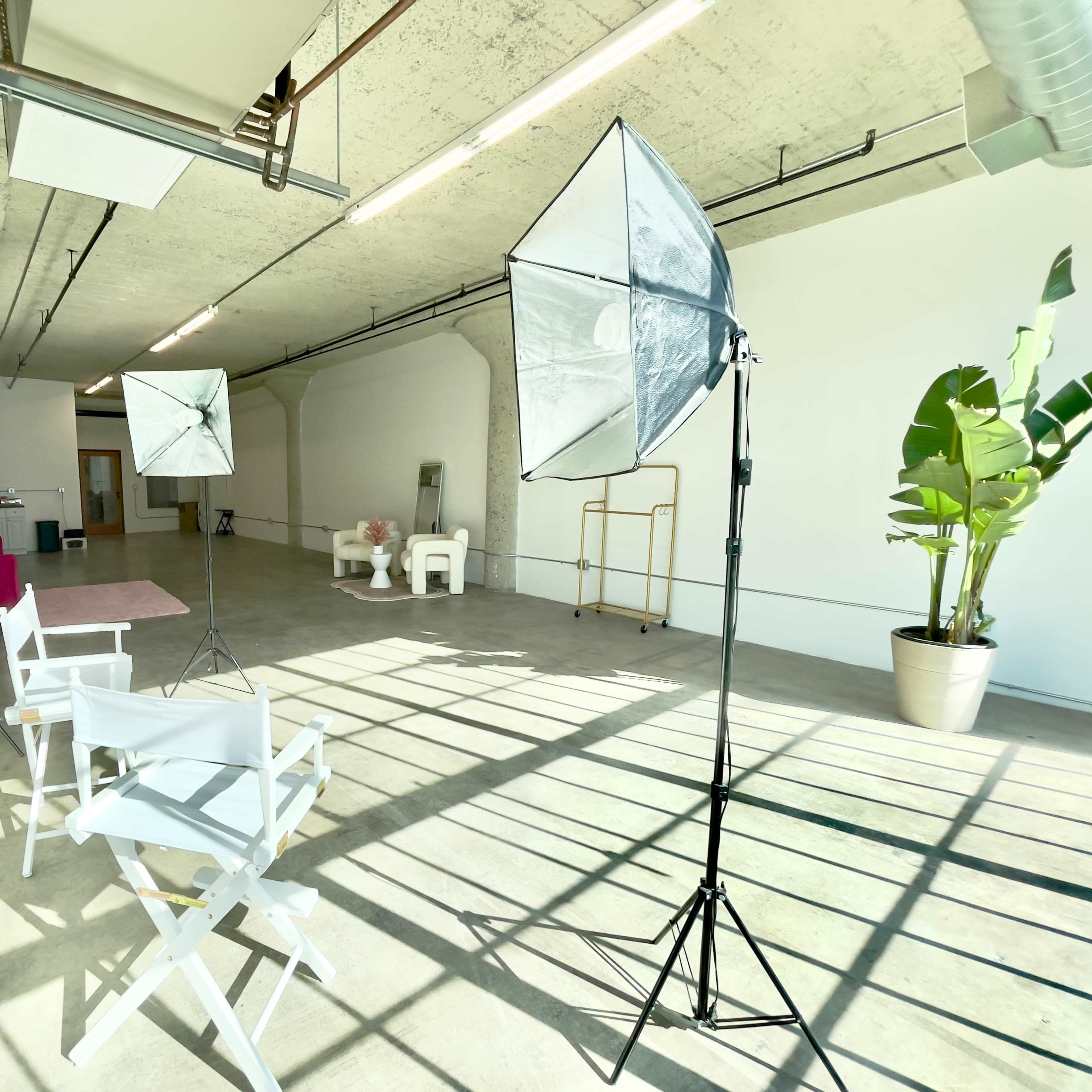 A spacious studio with concrete floors, a white backdrop, two light stands with softboxes, and two white director's chairs.