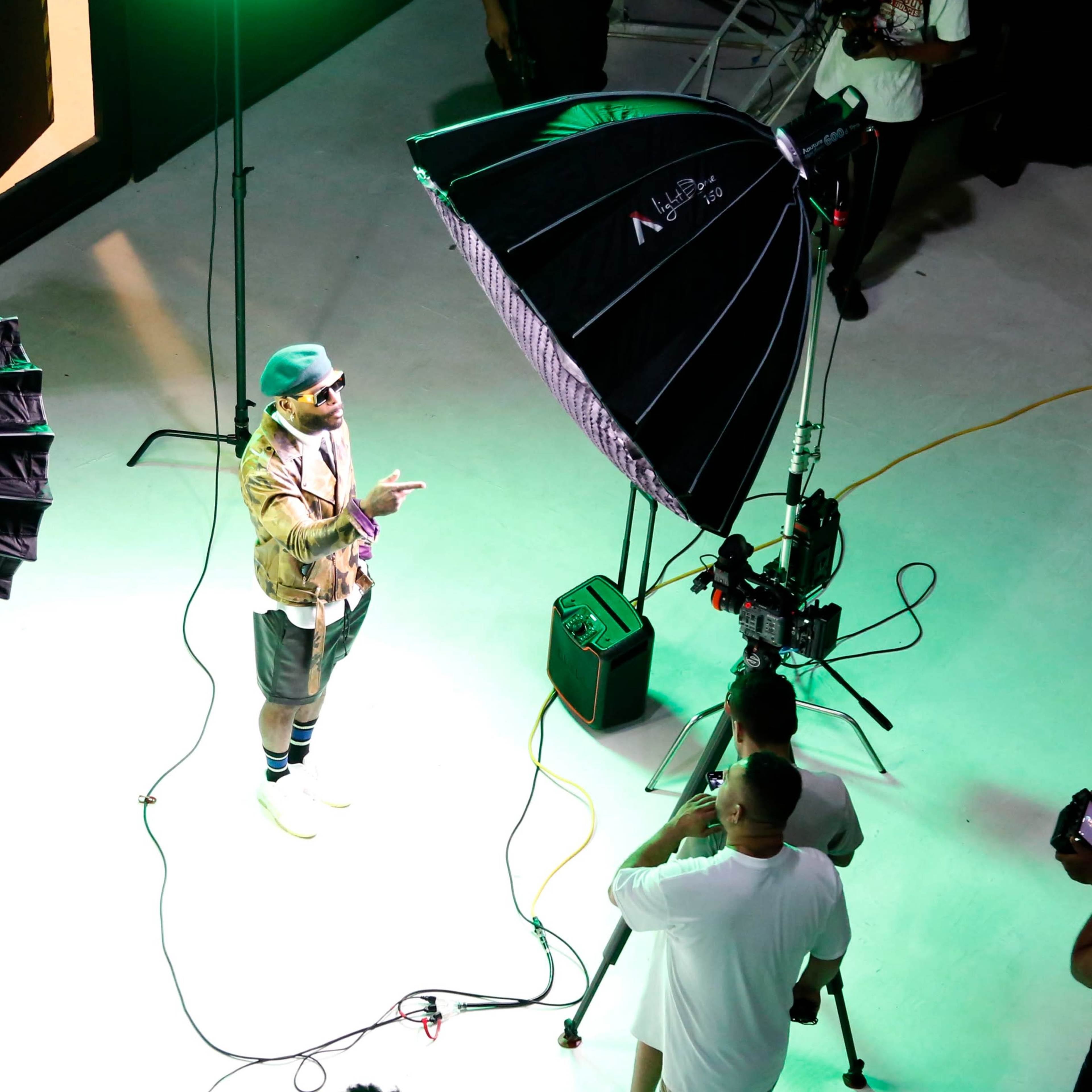 A male model stands on a lit platform surrounded by photography lighting equipment while a small crew prepares to take photos.