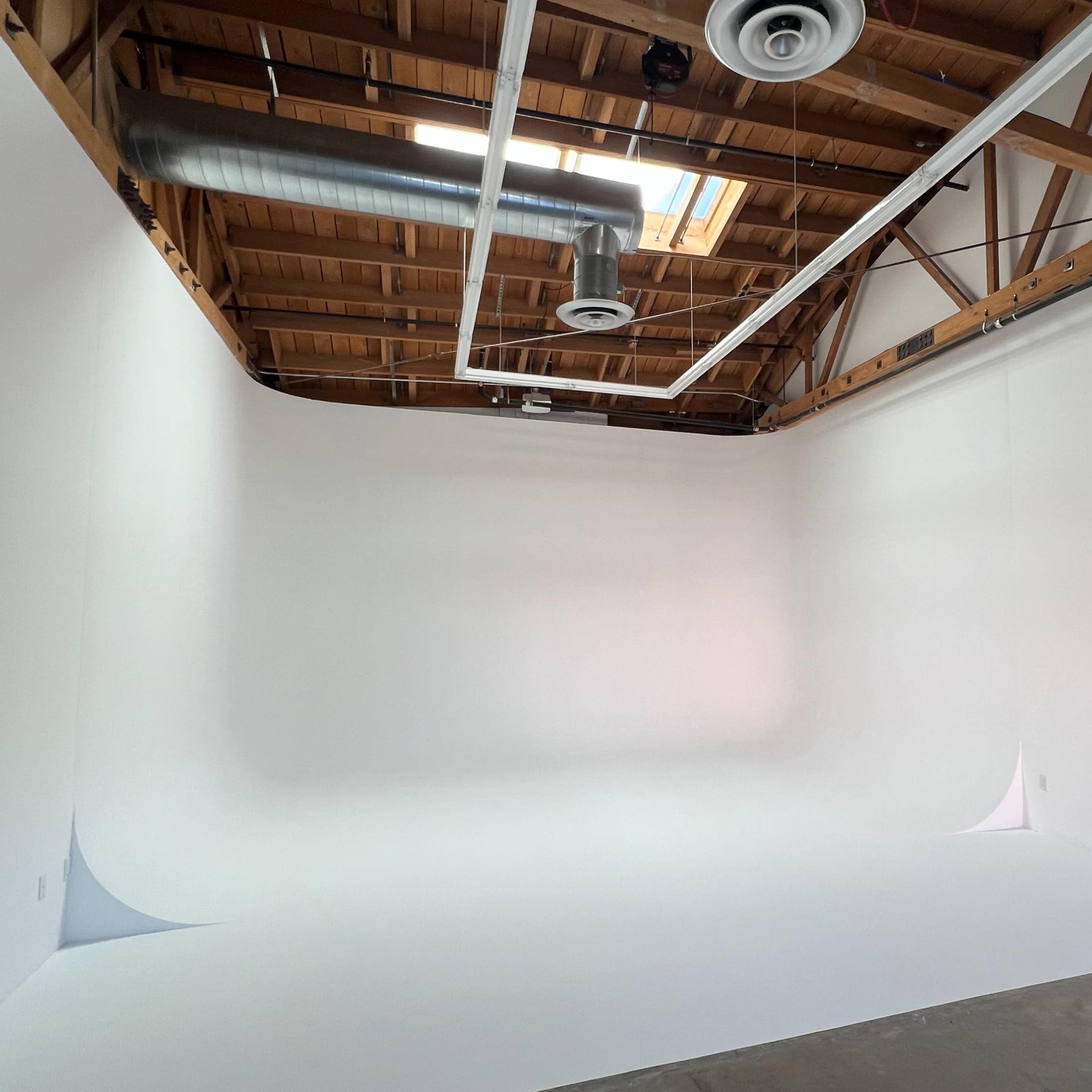 A spacious, empty studio with white walls, a smooth curved backdrop, and exposed wooden beams on the ceiling.