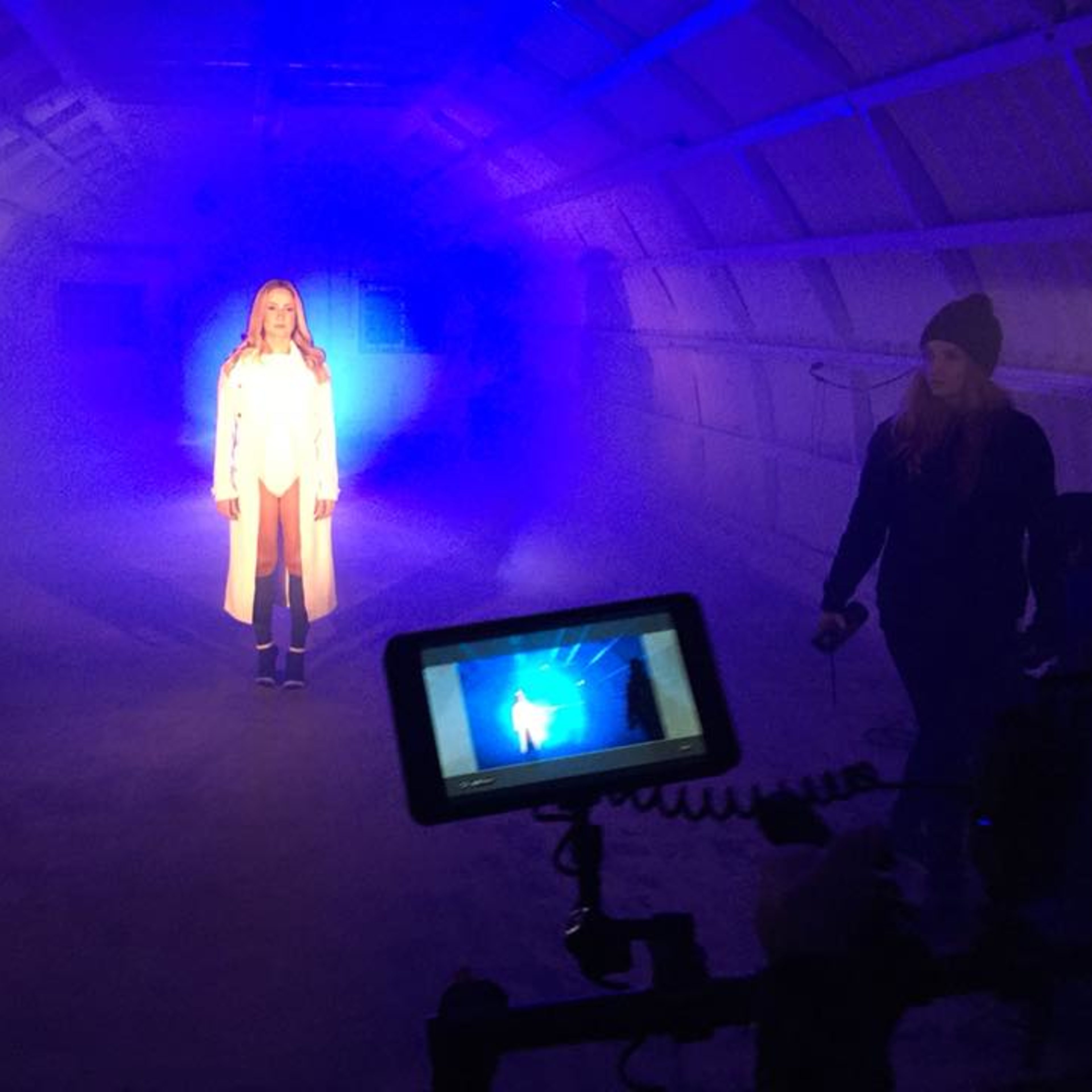 A woman stands in the center of a dimly lit, fog-filled space while a camera operator captures the scene on a monitor.