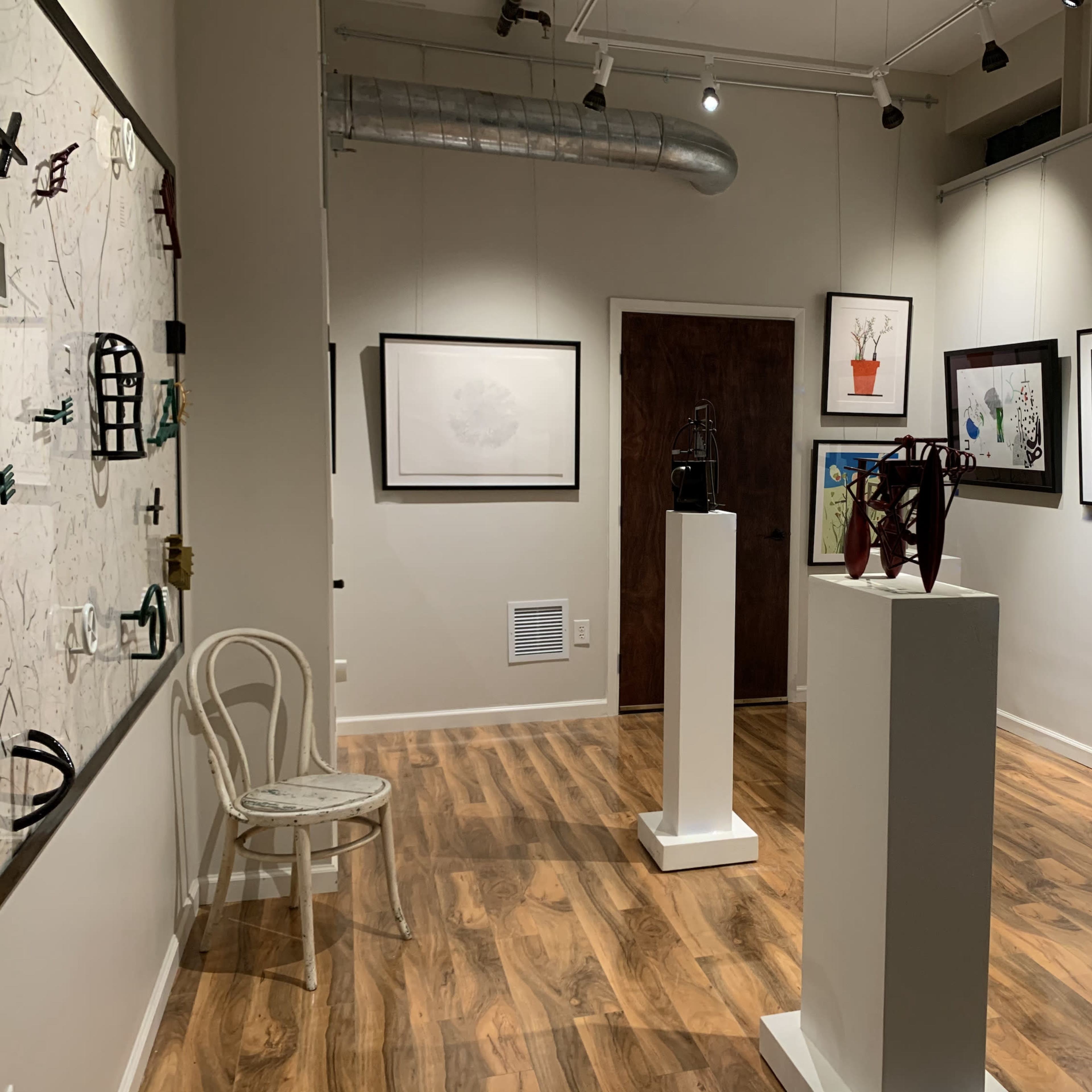 An art gallery with several framed artworks displayed on the walls and white pedestals featuring sculptures in a room with wooden flooring.