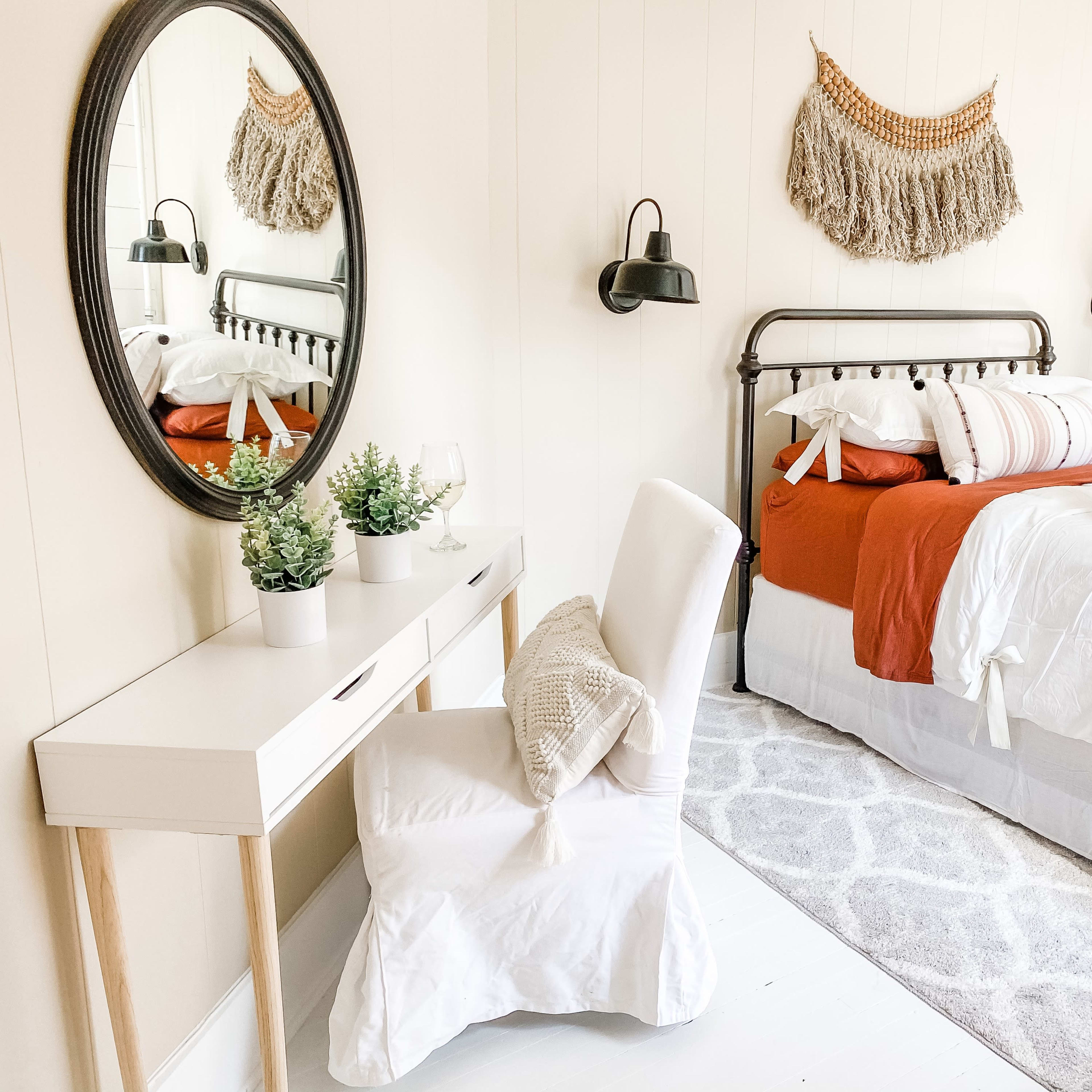 A cozy bedroom features a metal bed with red and white bedding, a round mirror above a white desk, and decorative plants on the desk and bedside.