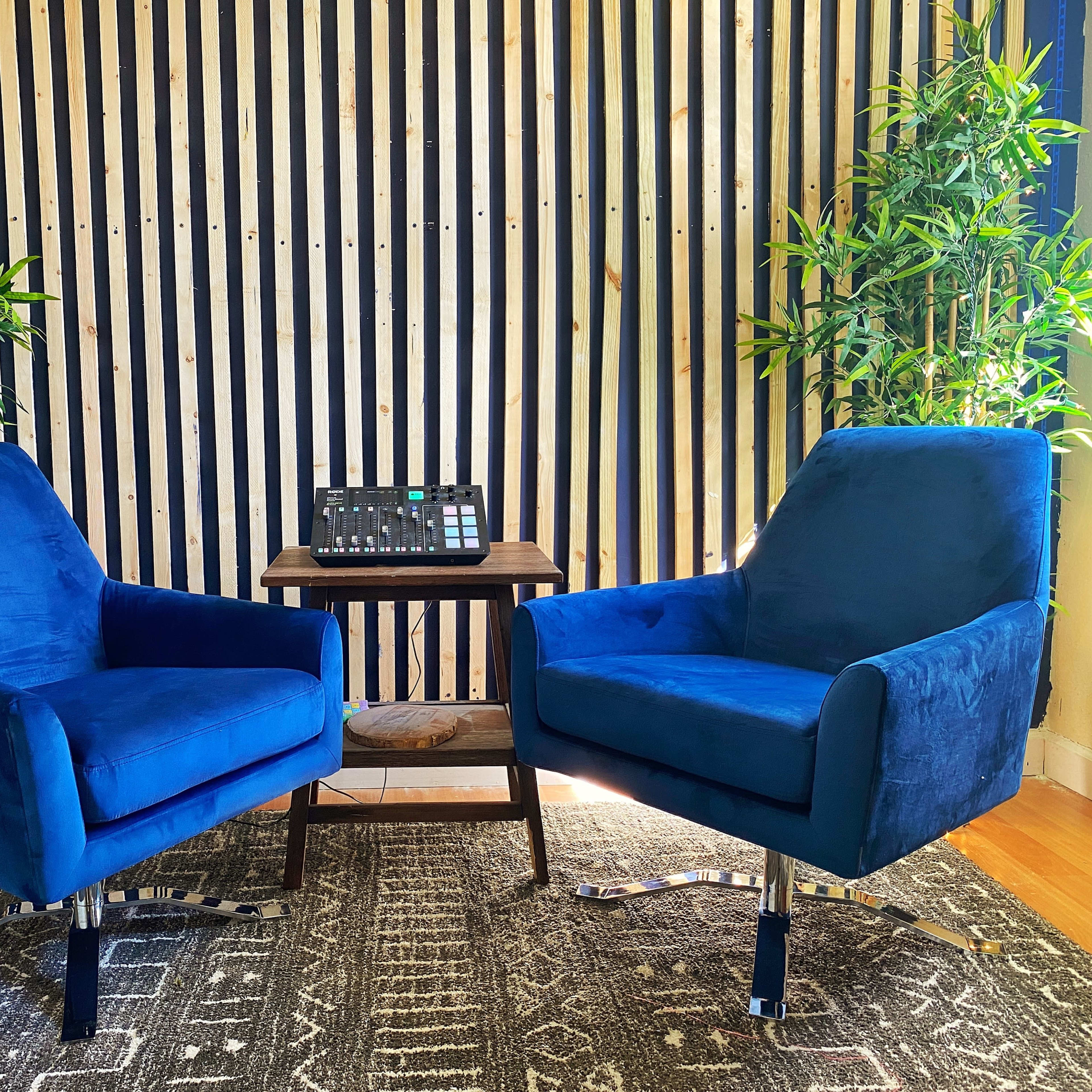 Two blue chairs positioned opposite each other with a small wooden table in between, set against a striped wooden wall and flanked by green plants.