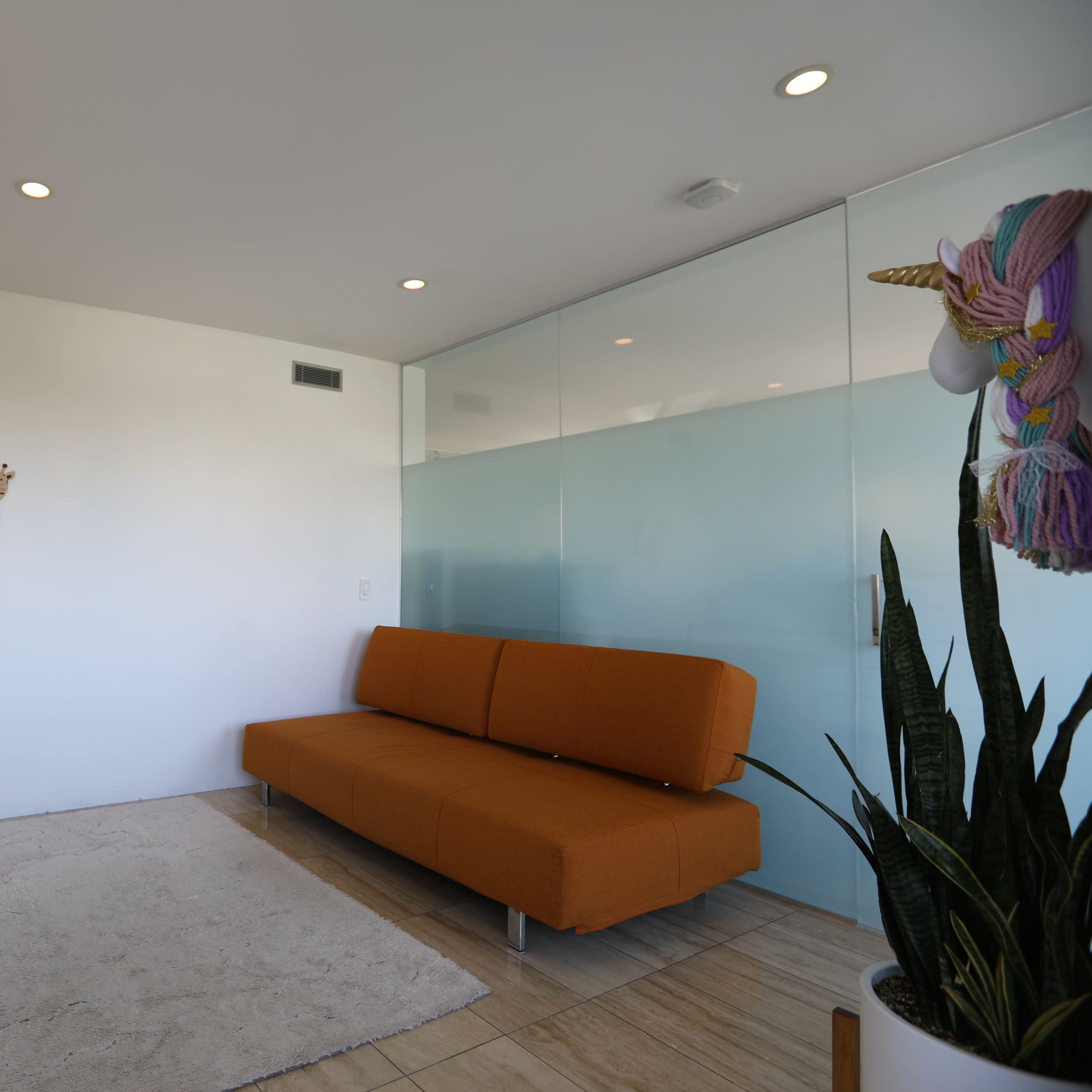 A modern, minimalist living space with an orange sofa, a rug on tiled flooring, and decorative wall hangings near a glass partition.