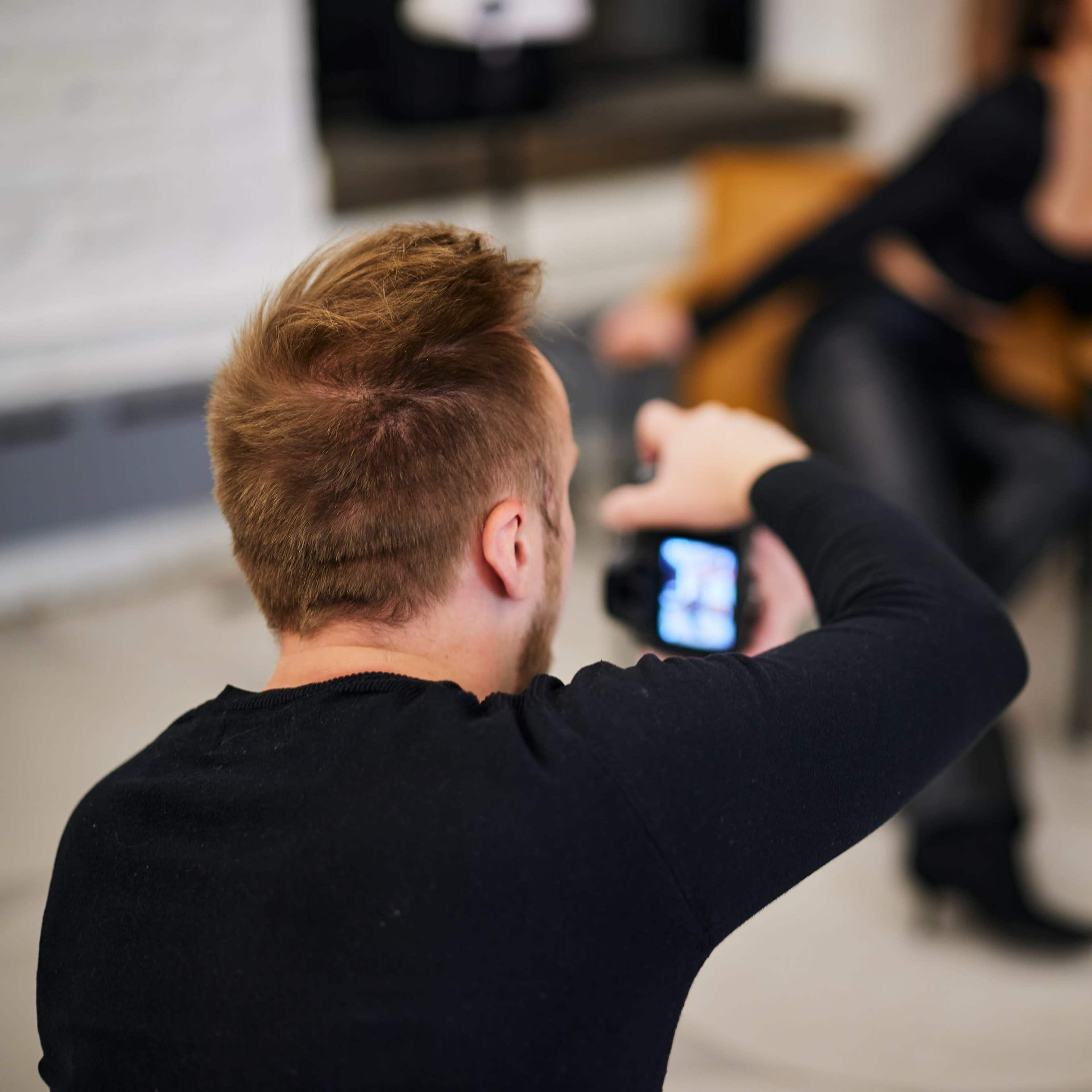 A photographer captures a model posed in a chair while focusing on his camera in a well-lit indoor setting.