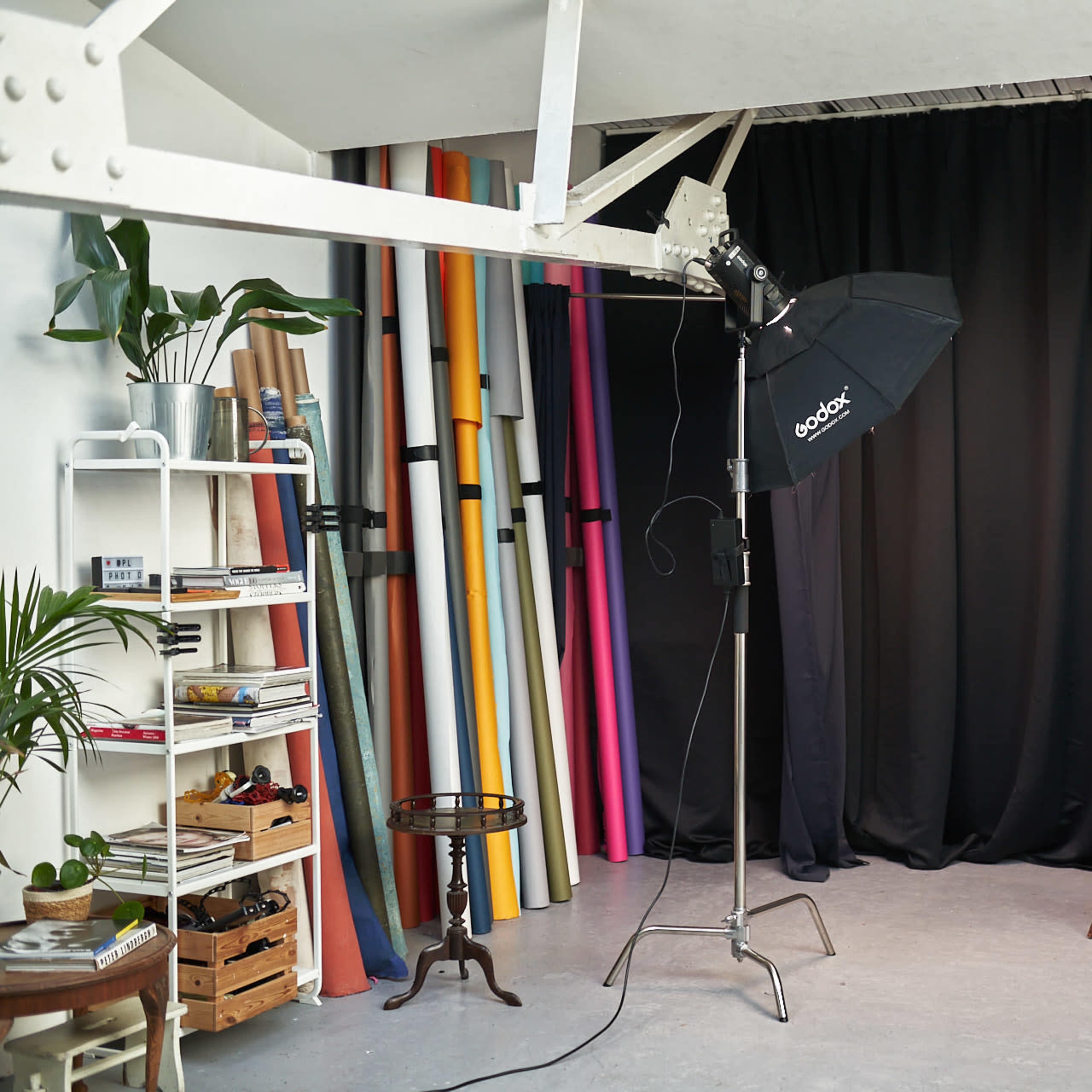 A photography studio with a black backdrop, a lighting setup, colorful rolls of paper, a chair, and shelves displaying various items.