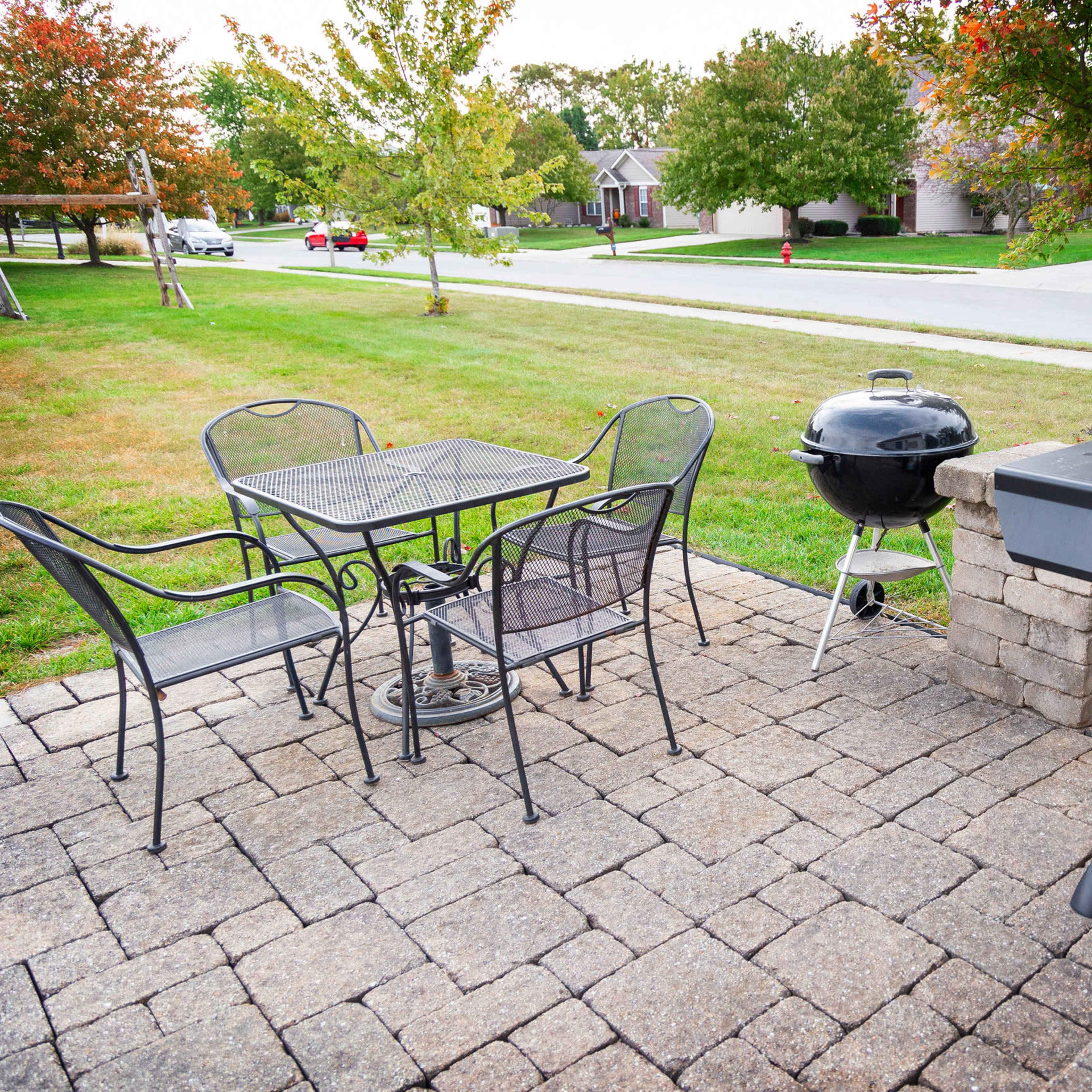 A patio area with a black gas grill, a charcoal grill, a metal table with four chairs, and landscaped greenery in the background.