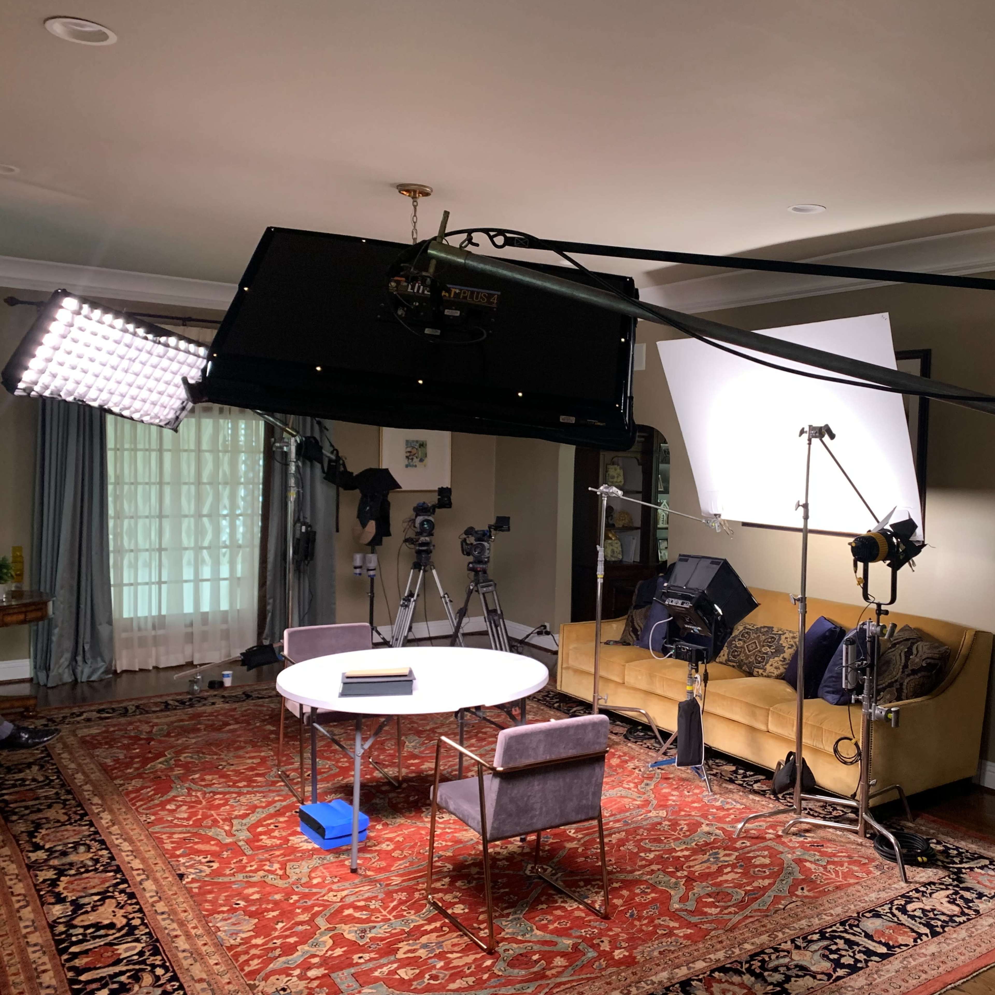 A television studio setup features a round table, two chairs, and multiple cameras and lights arranged in a living room with a patterned rug.