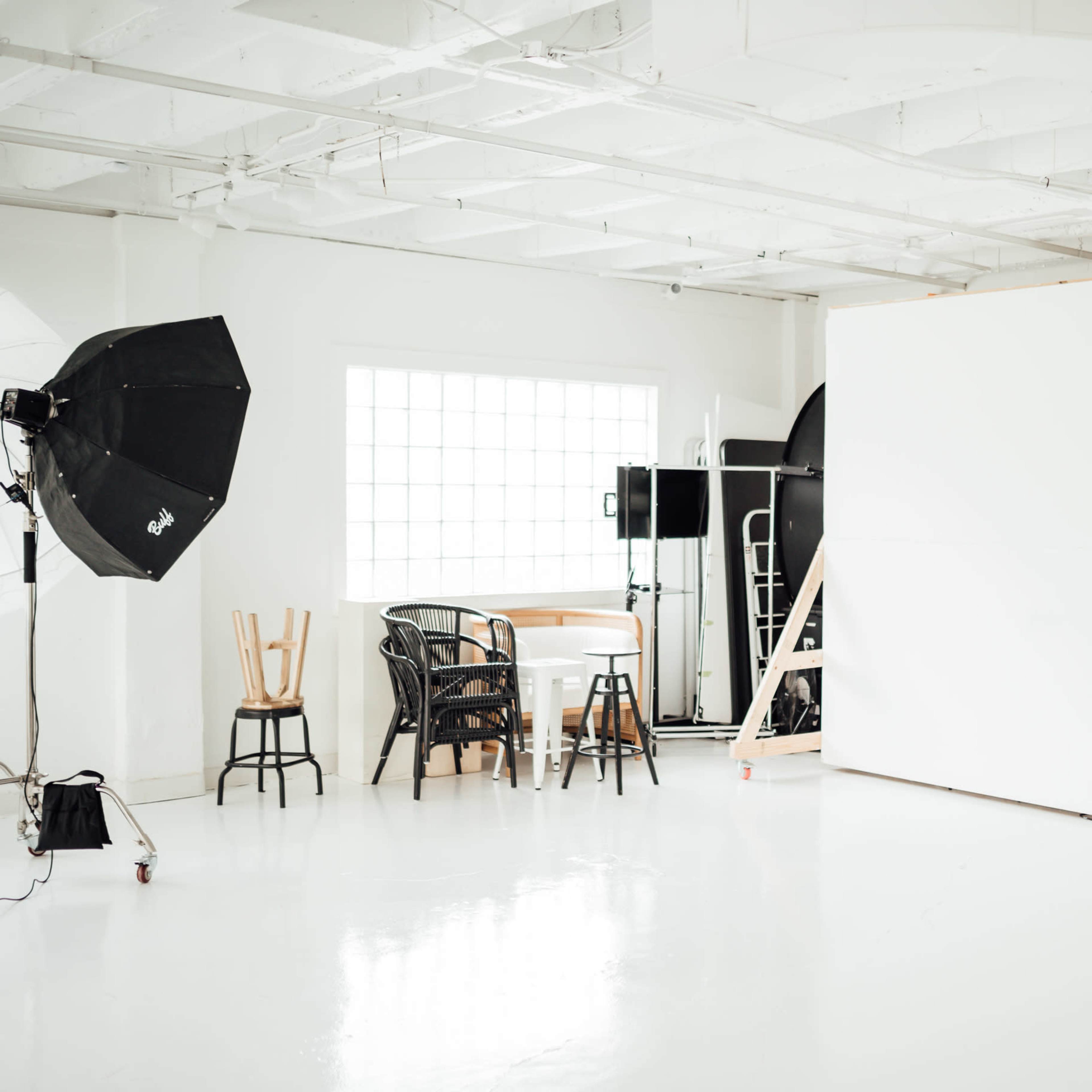 A photography studio with a large white backdrop, a table and chairs, and various lighting equipment.