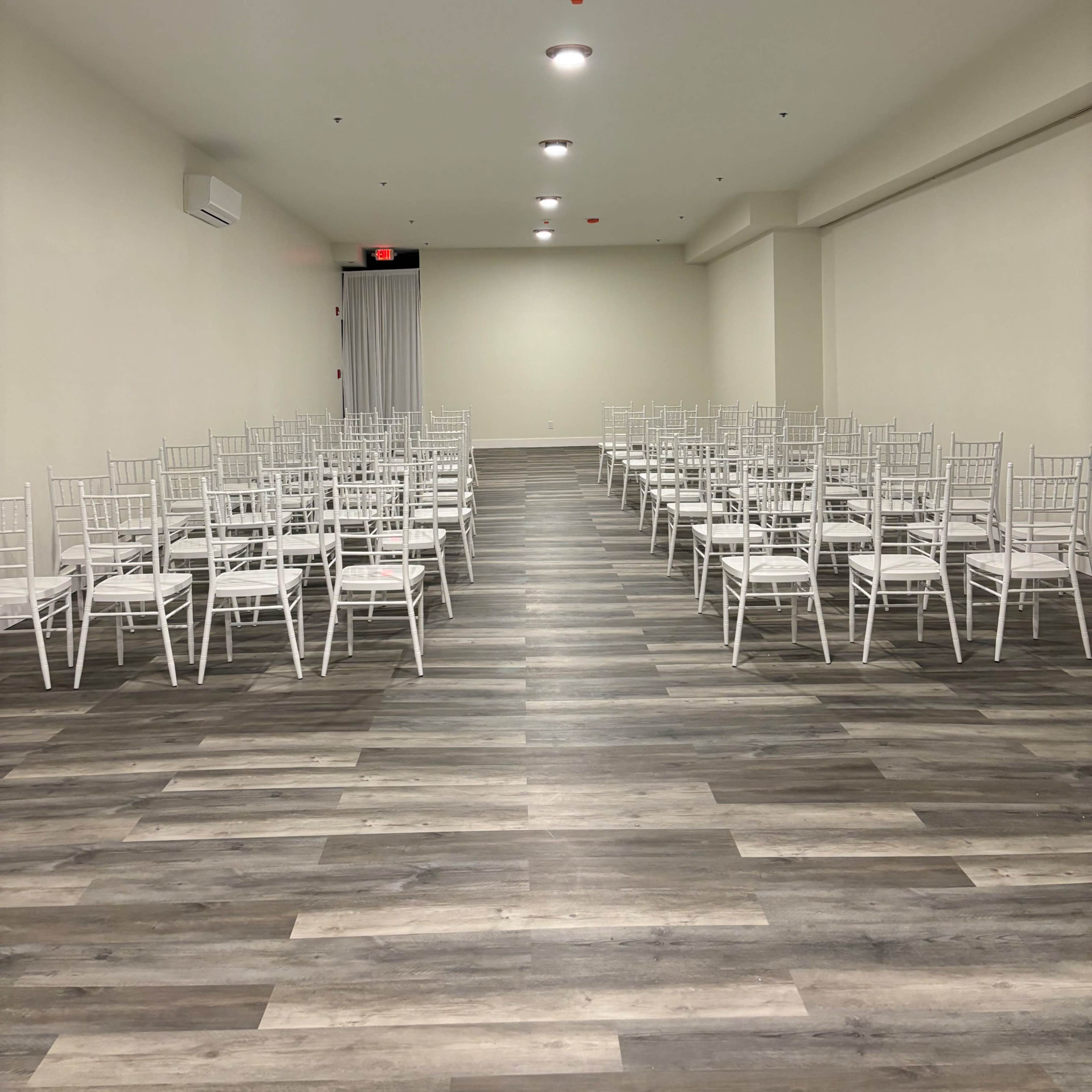 A spacious room with rows of white chiavari chairs arranged on a wooden floor.