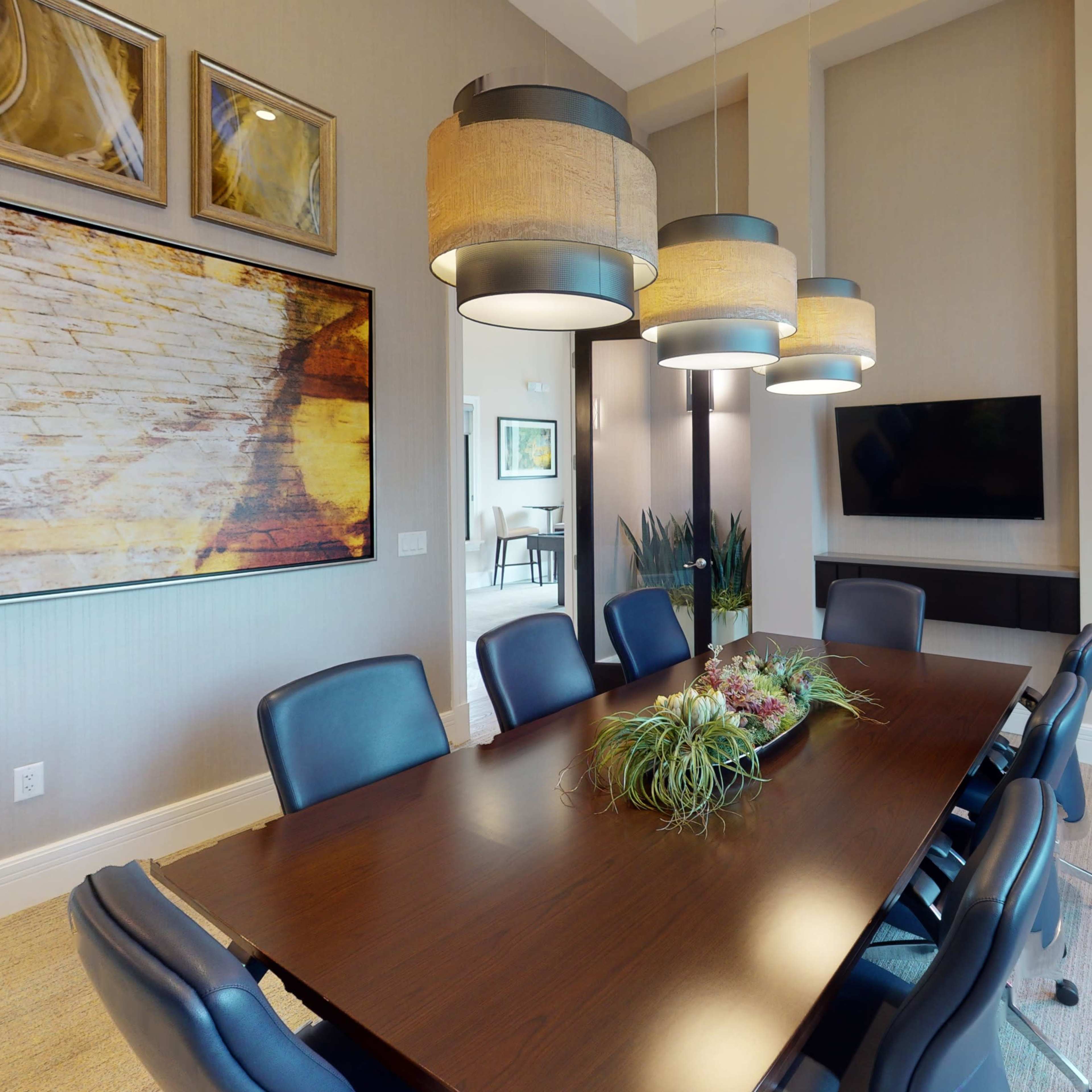 A modern conference room with a large wooden table surrounded by black chairs, featuring pendant lights, artwork on the walls, and a television.