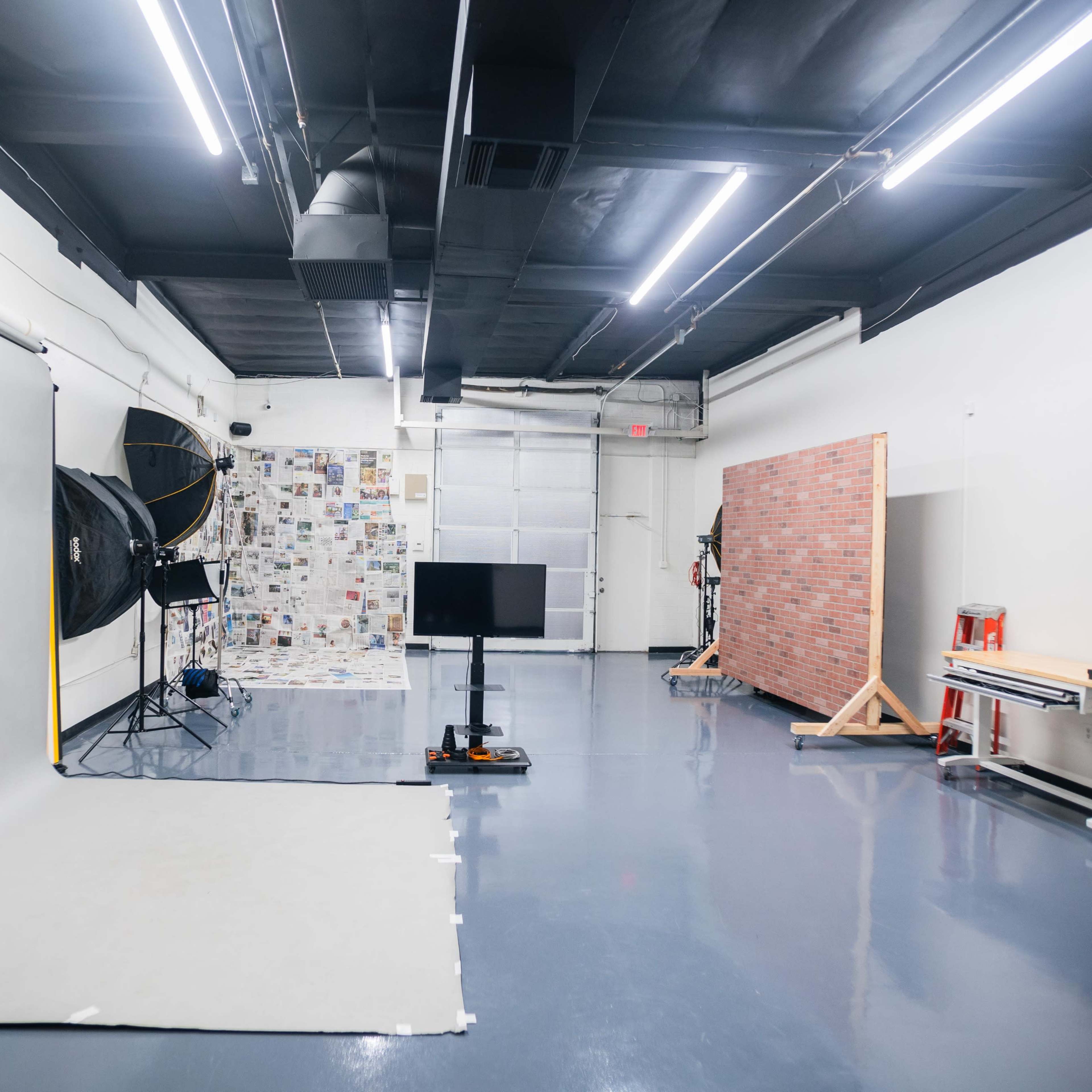 A photography studio with a seamless backdrop, a large softbox, a television monitor on a stand, and various props against a white wall.