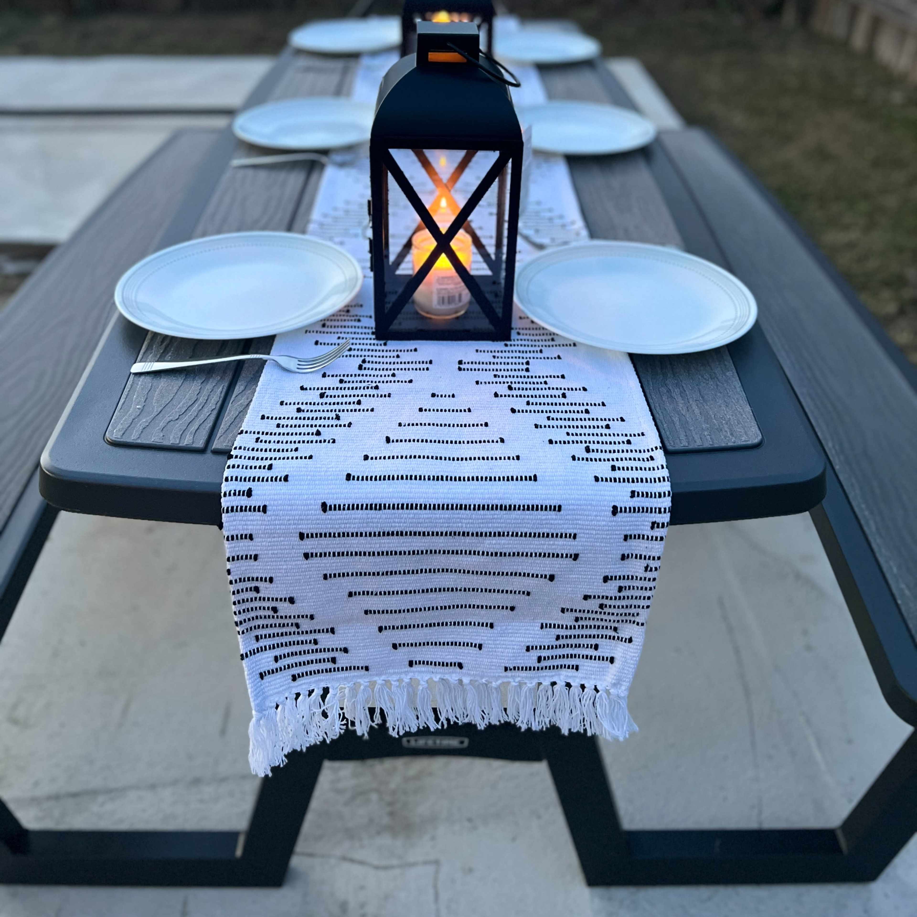 A picnic table set for a meal features white plates and lanterns on a patterned table runner, with a hammock visible in the background.