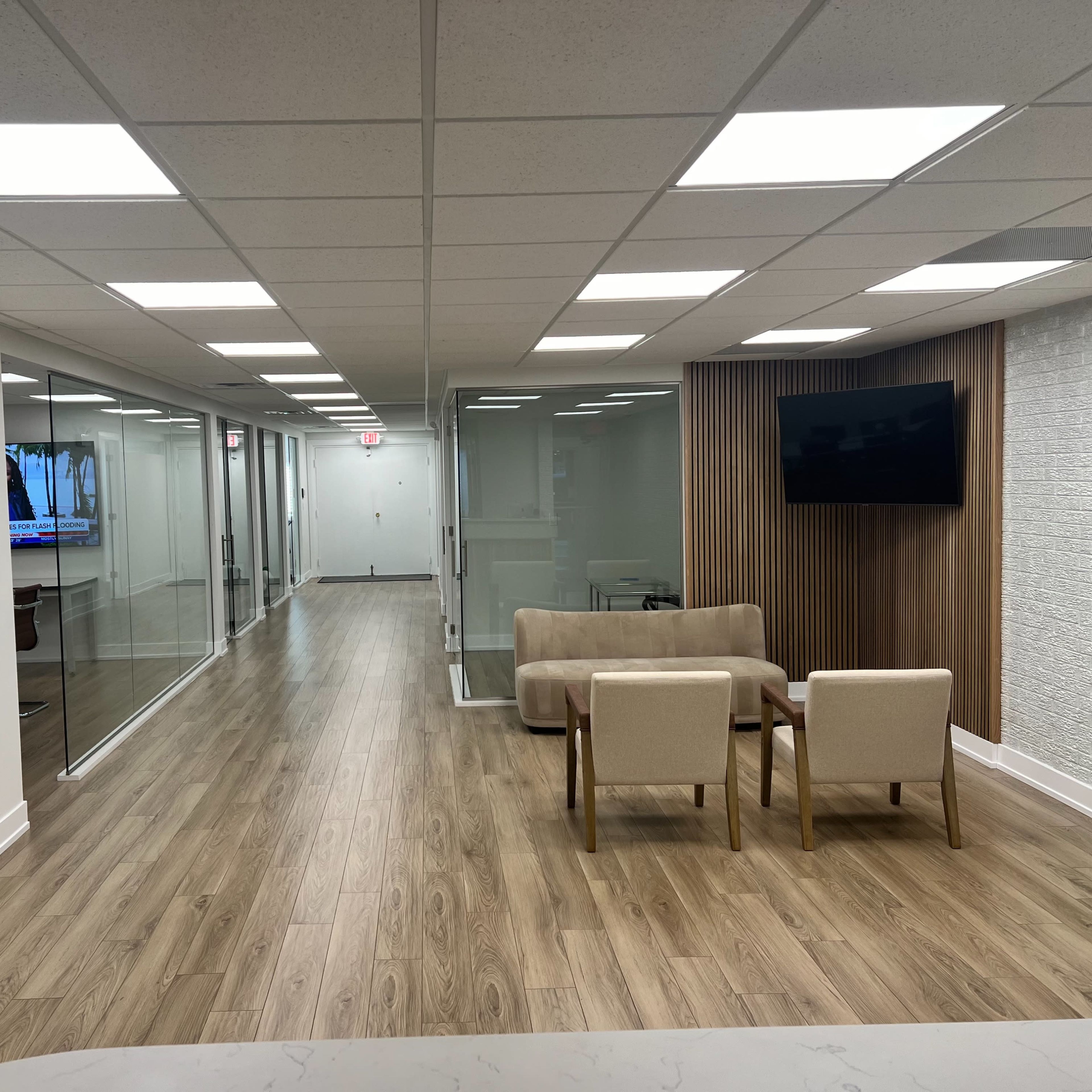 A modern office hallway with glass-walled conference rooms and a seating area featuring two chairs and a sofa.