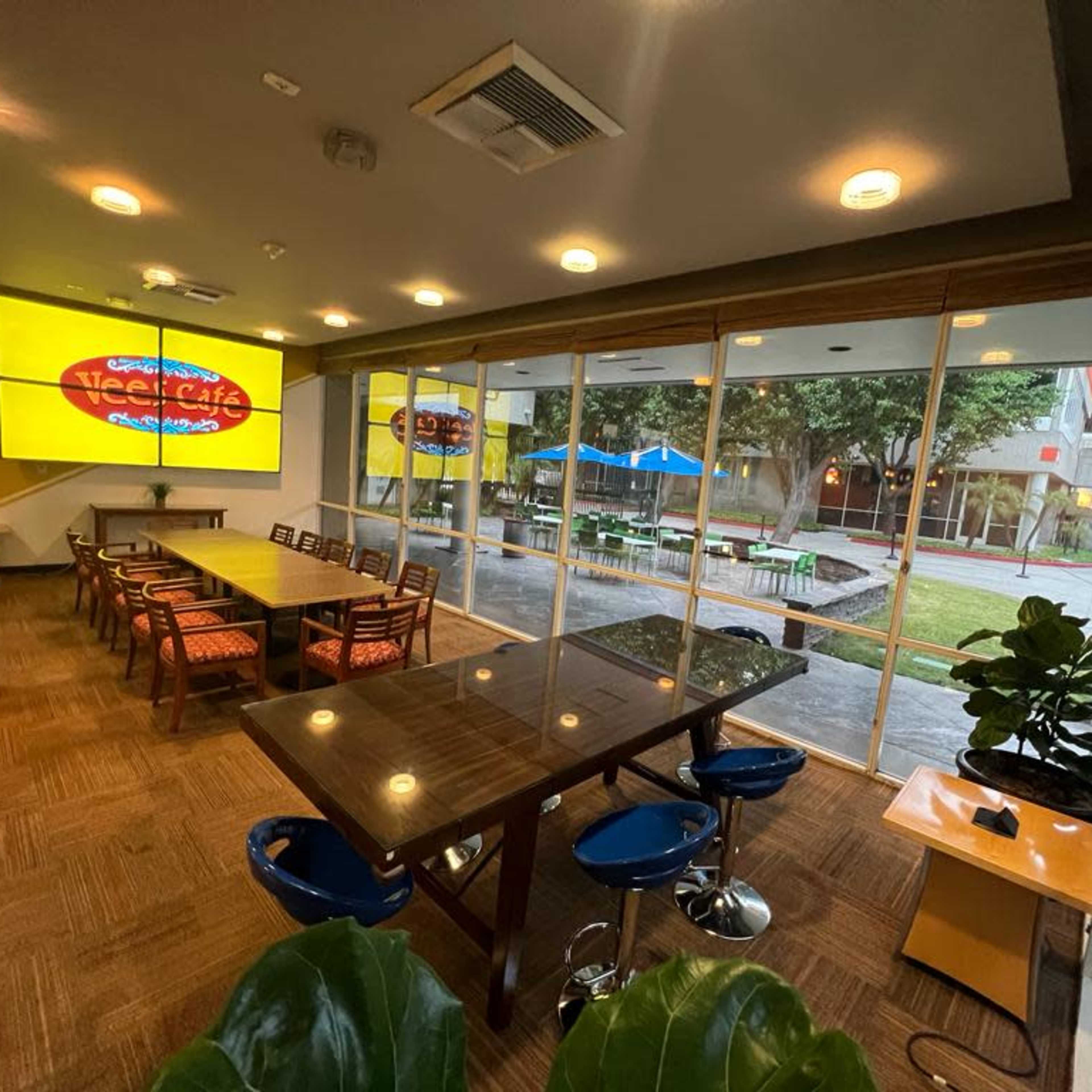 A spacious indoor café area with a large table and several smaller tables, large windows overlooking an outdoor space with seating, and a bright screen displaying the café's name.
