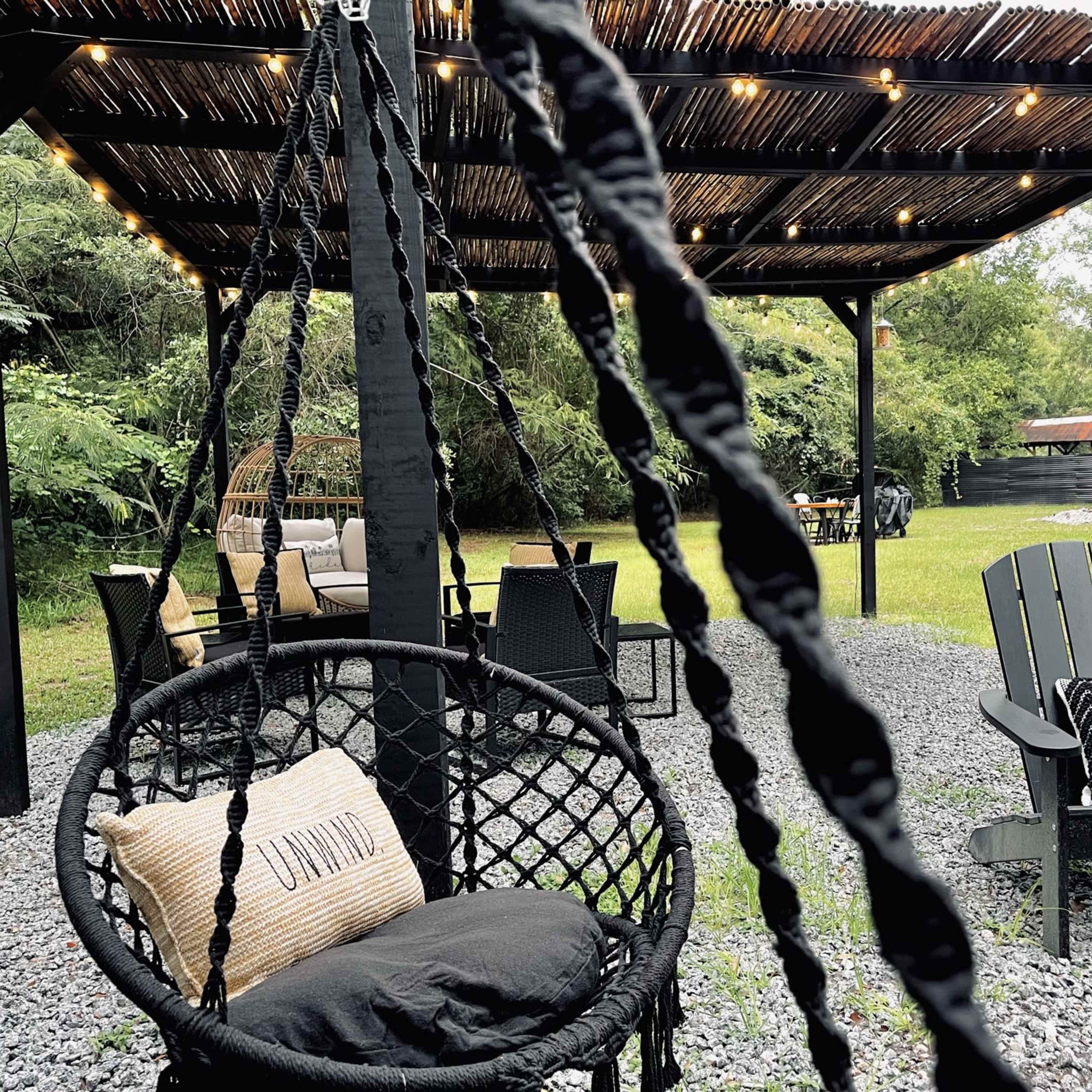 A shaded outdoor seating area features hanging chairs, a cozy couch, and string lights above, surrounded by greenery and gravel.