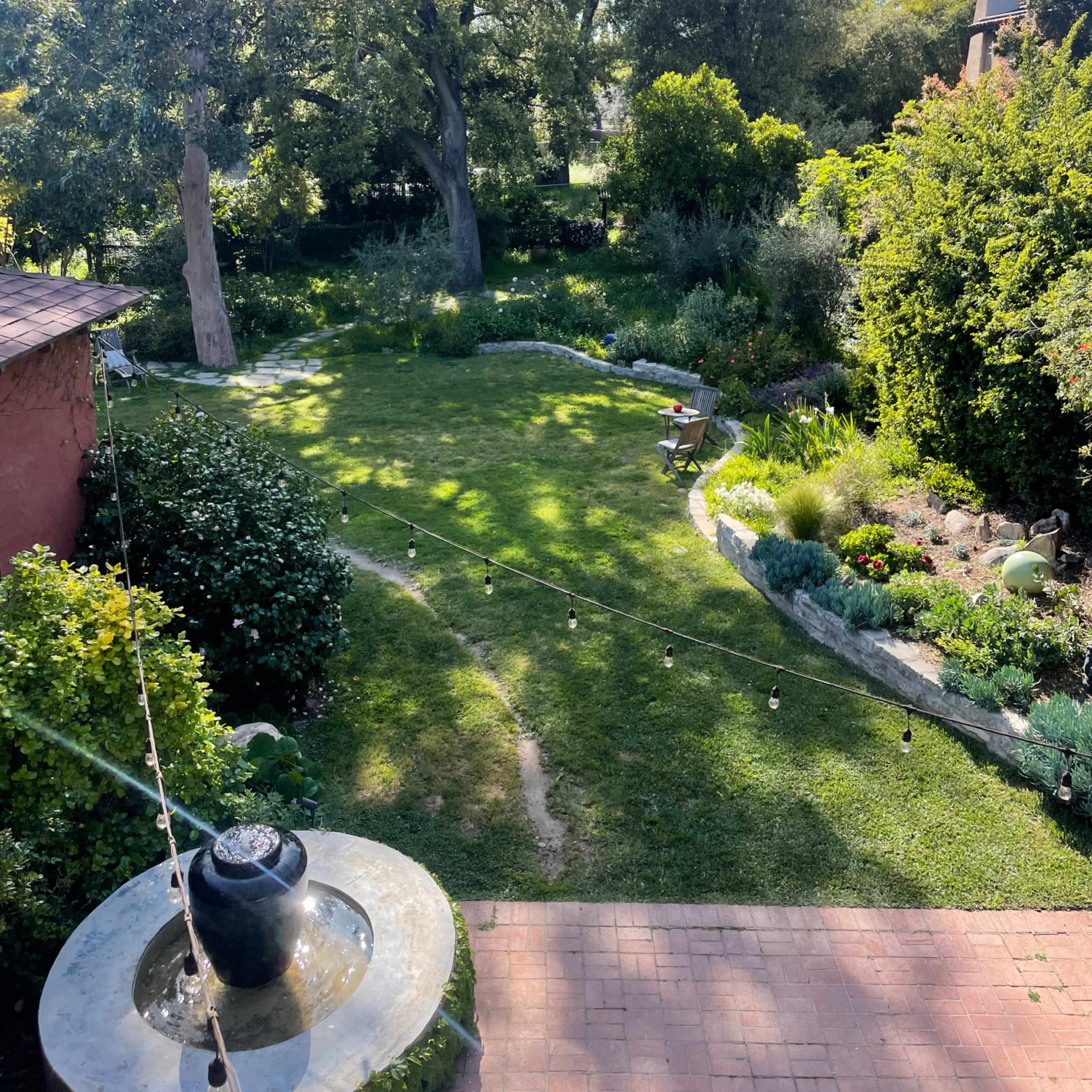 A well-maintained garden with a lawn, bordered by various plants and trees, and featuring a stone fountain in the foreground.
