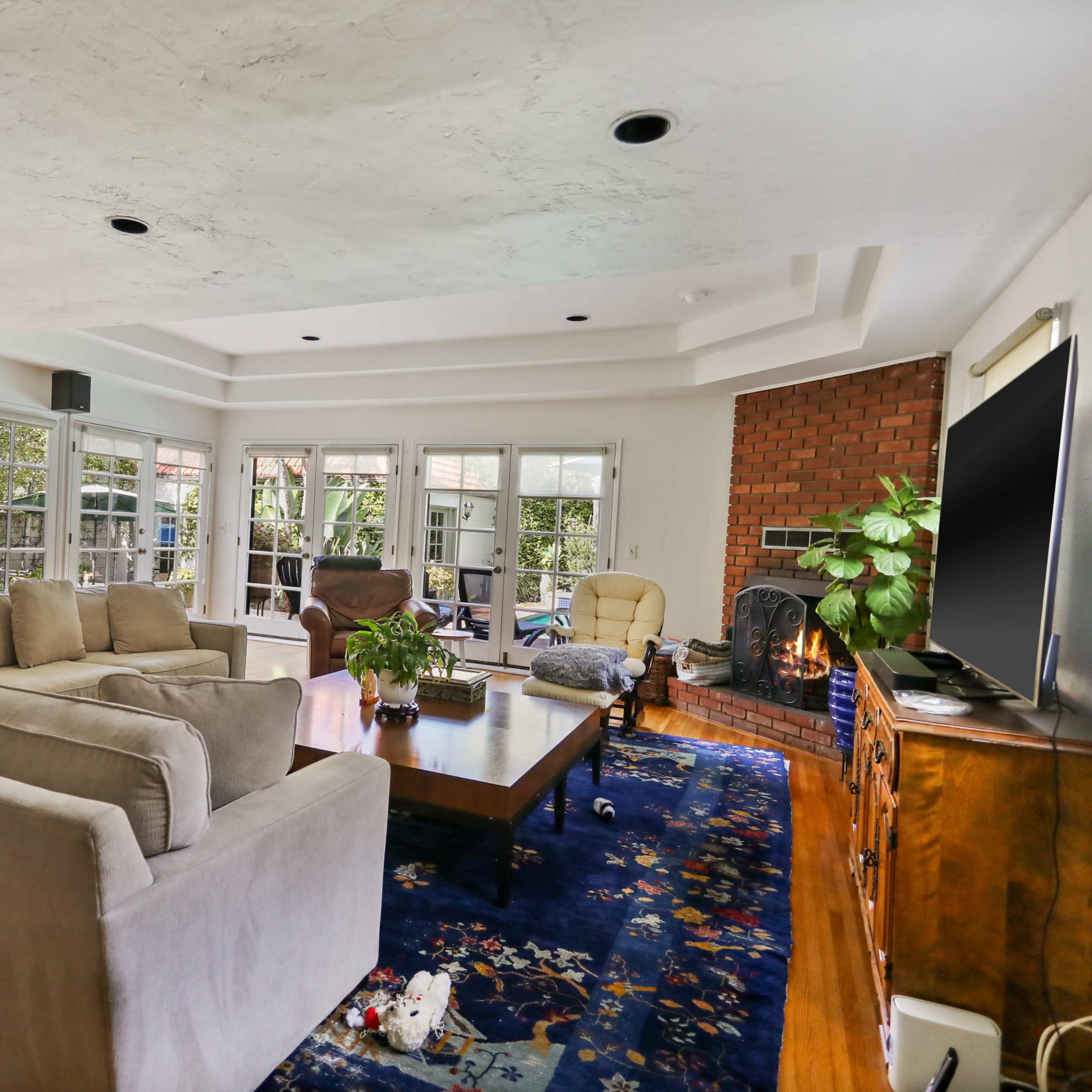 A spacious living room features a sofa set, a wooden coffee table, and a brick fireplace, with large windows providing views of the outdoors.
