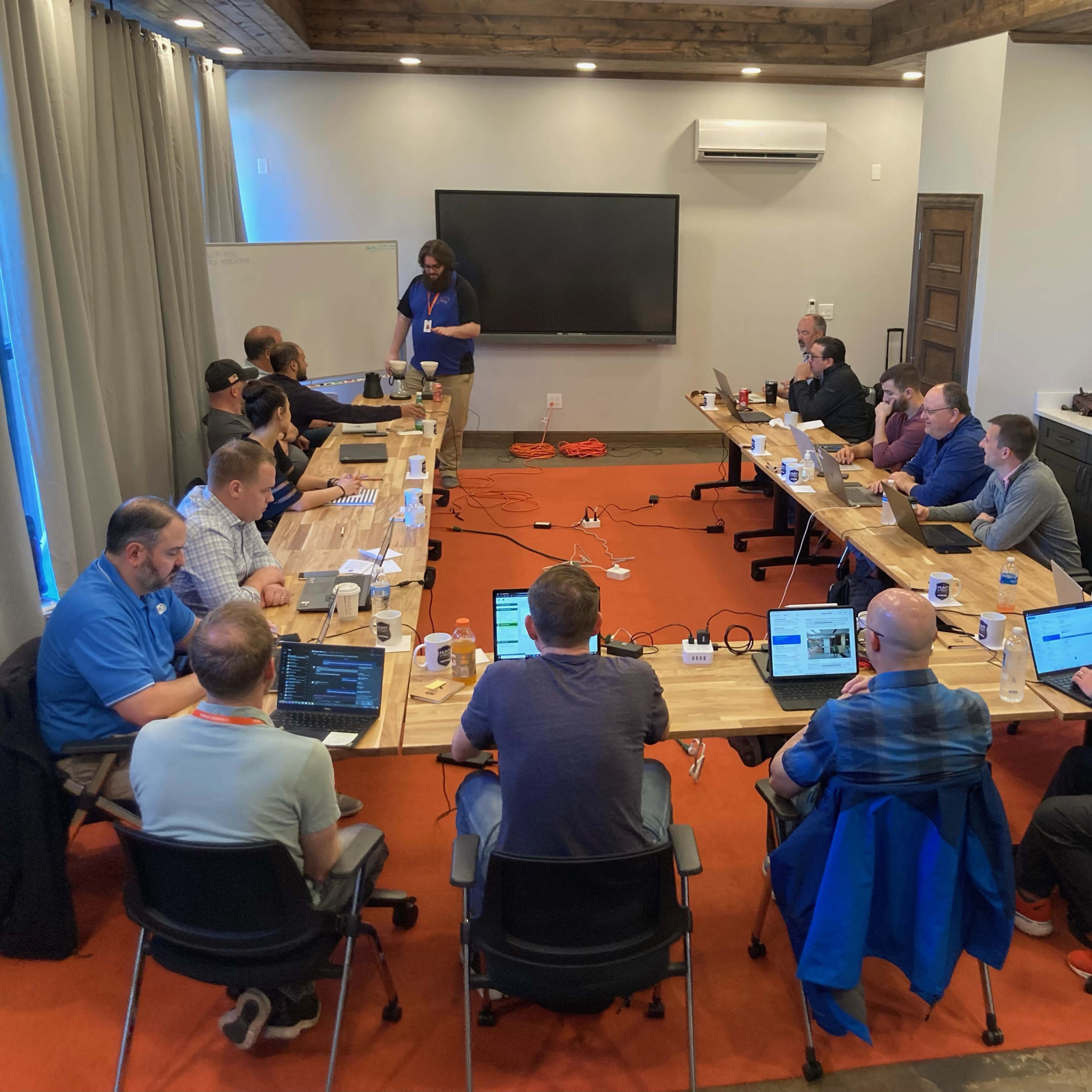 A group of professionals is seated around a long table in a conference room, actively engaged in a meeting with laptops and notes in front of them.