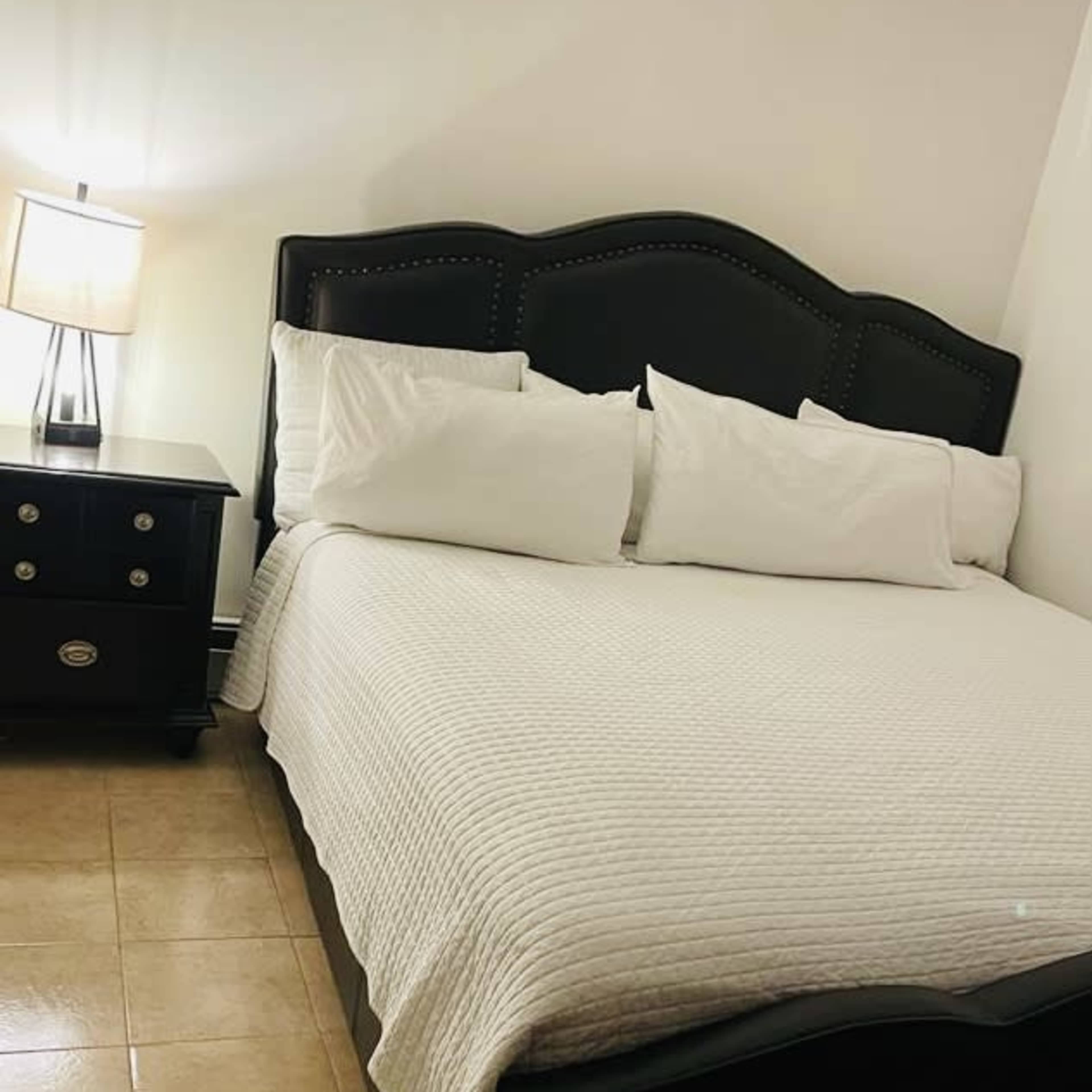 A neatly made bed with white pillows and a light-colored quilt, positioned next to a lamp and a dark bedside table.