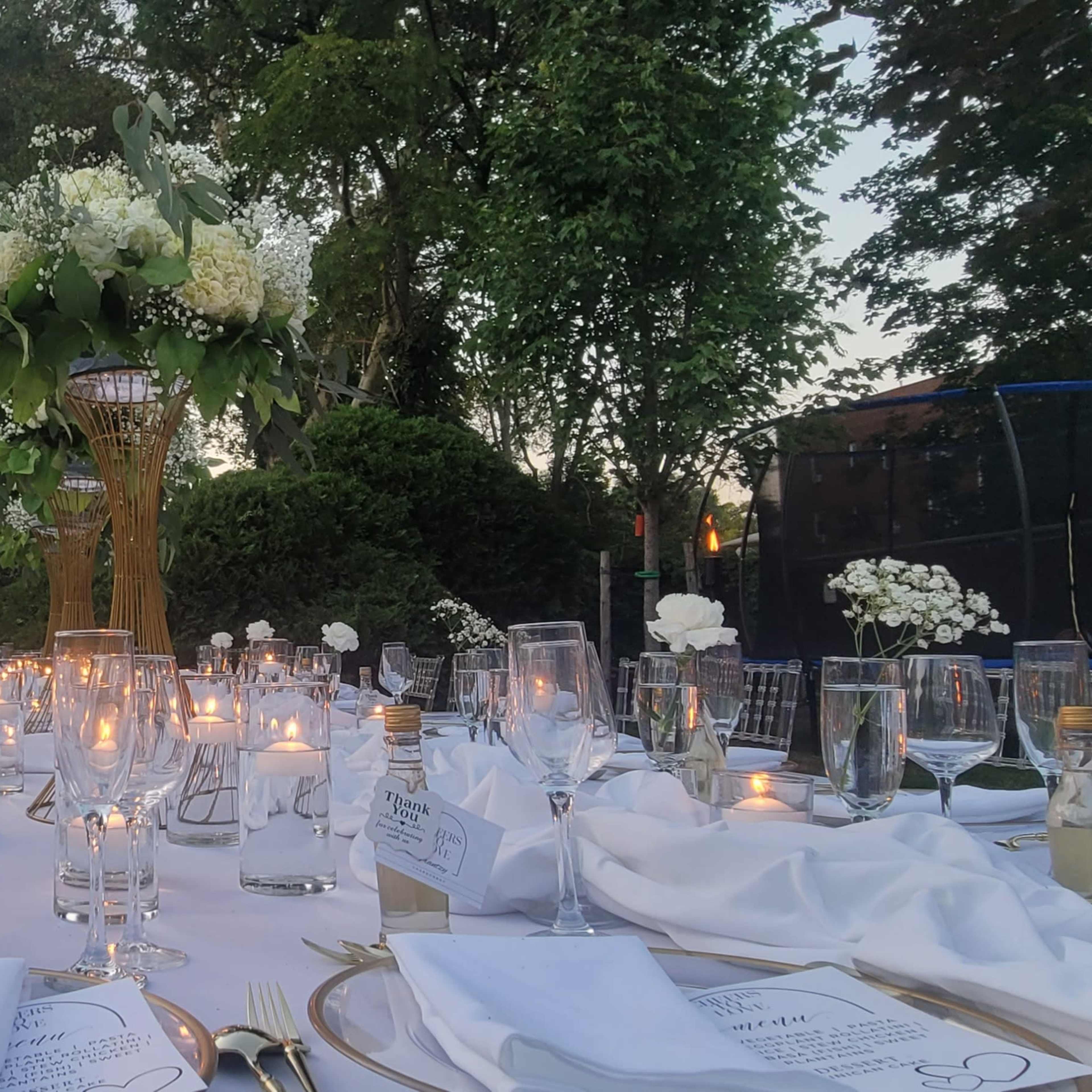 A beautifully arranged outdoor dining table is set with elegant white linens, glassware, and floral centerpieces, illuminated by soft candlelight.