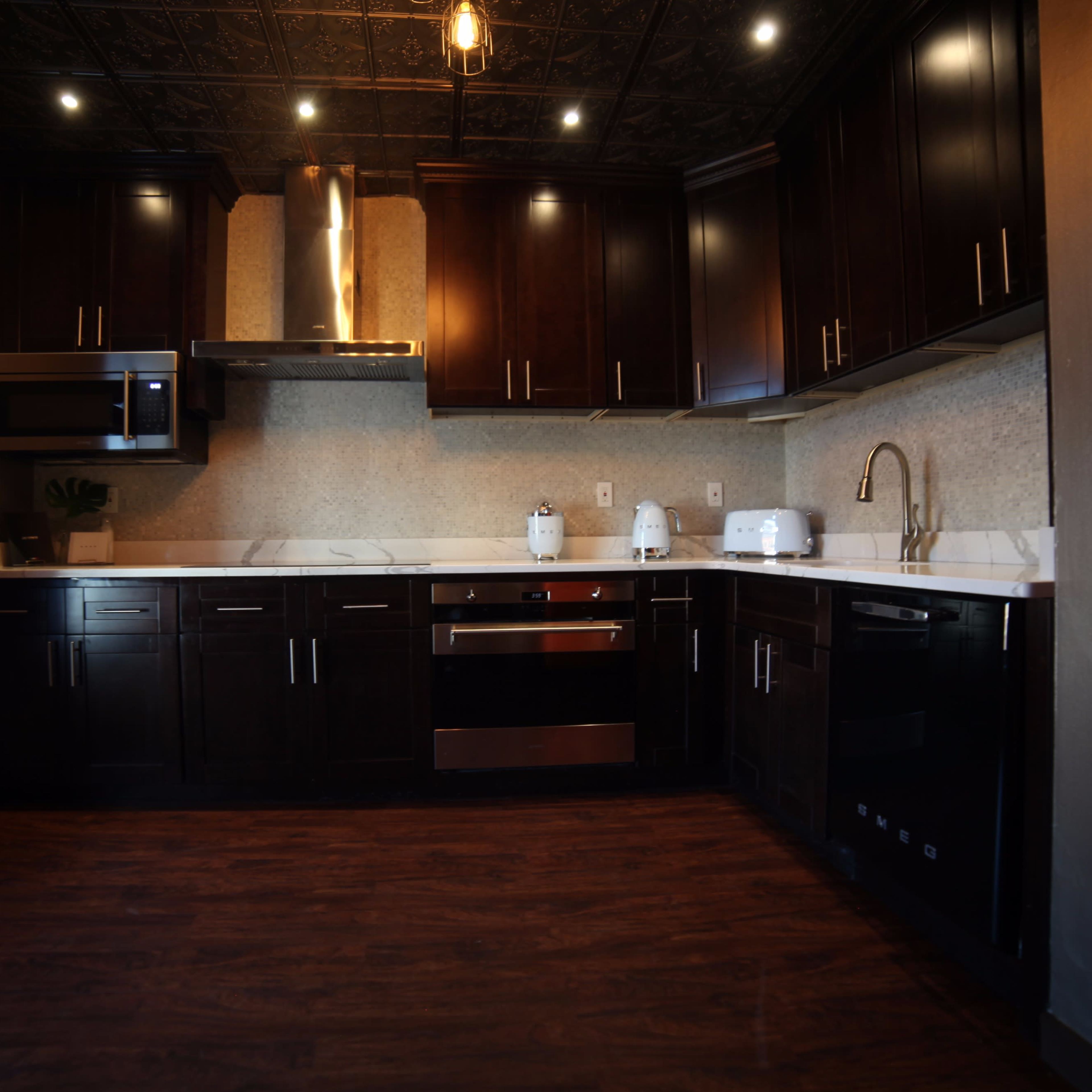 A modern kitchen with dark wooden cabinets, stainless steel appliances, and a white countertop illuminated by overhead lights.