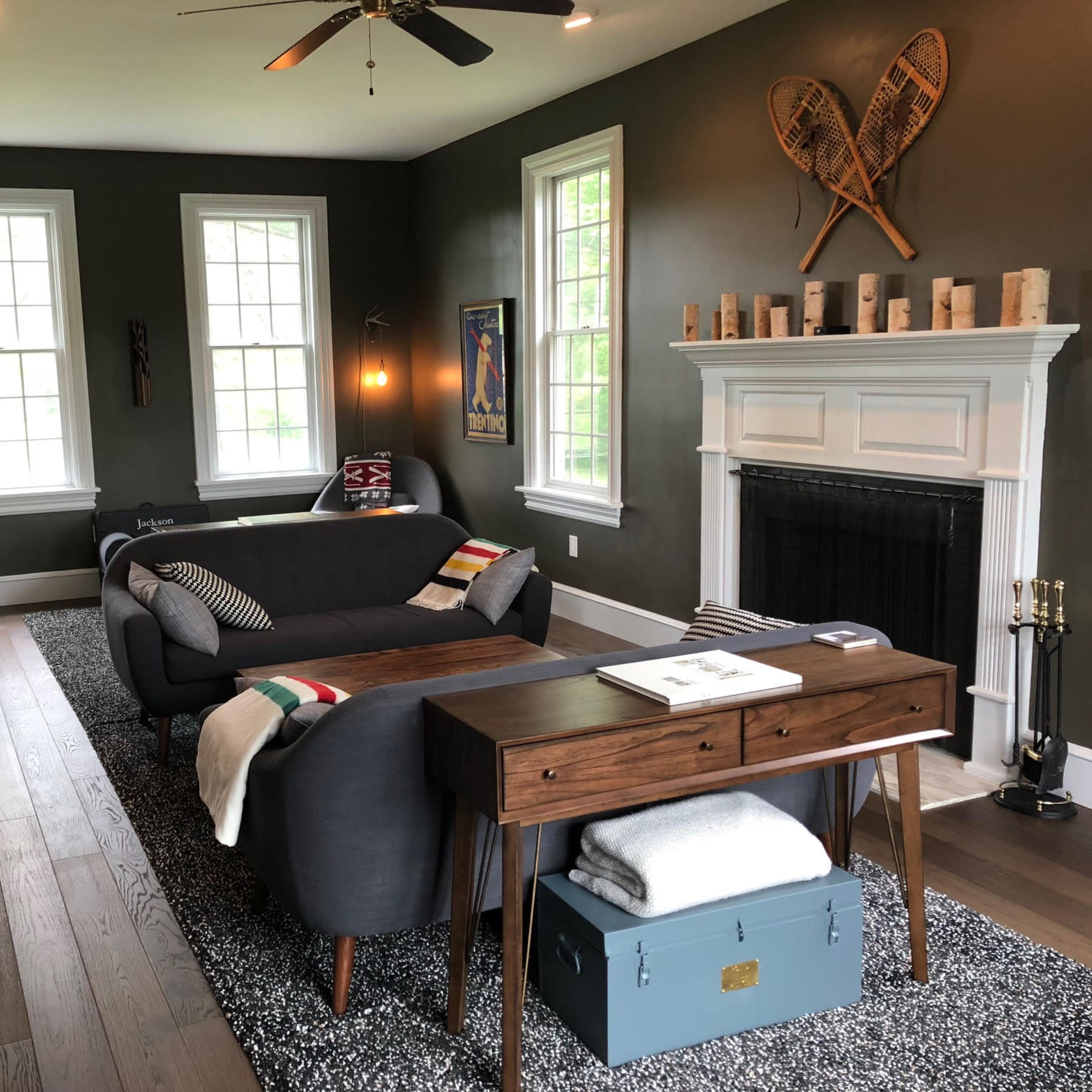 A modern living room featuring two gray sofas, a wooden table, a fireplace, and decorative wall art in a space with dark green walls and large windows.