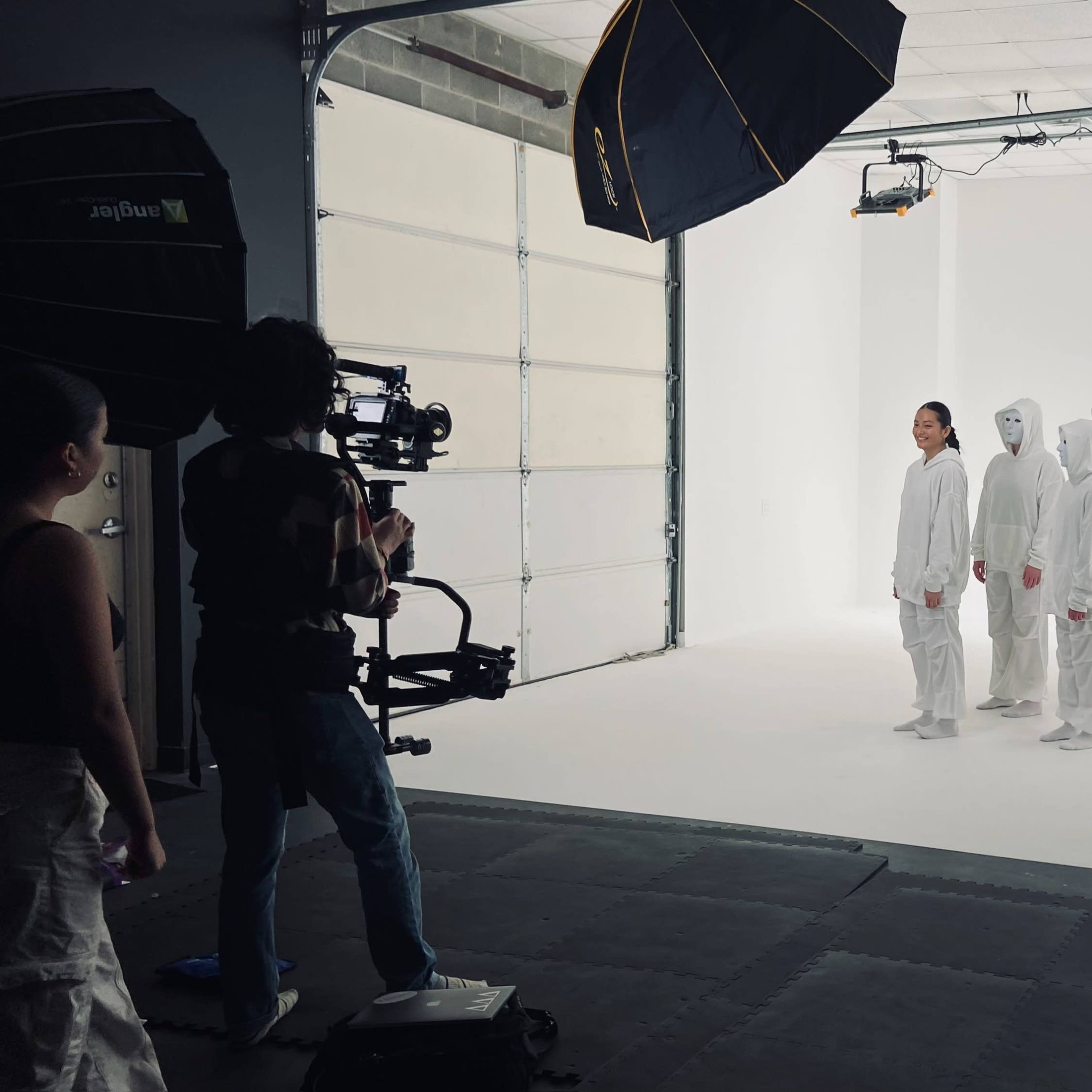 A group of individuals dressed in white clothing stands in a line on a white backdrop while a person operates a camera nearby.