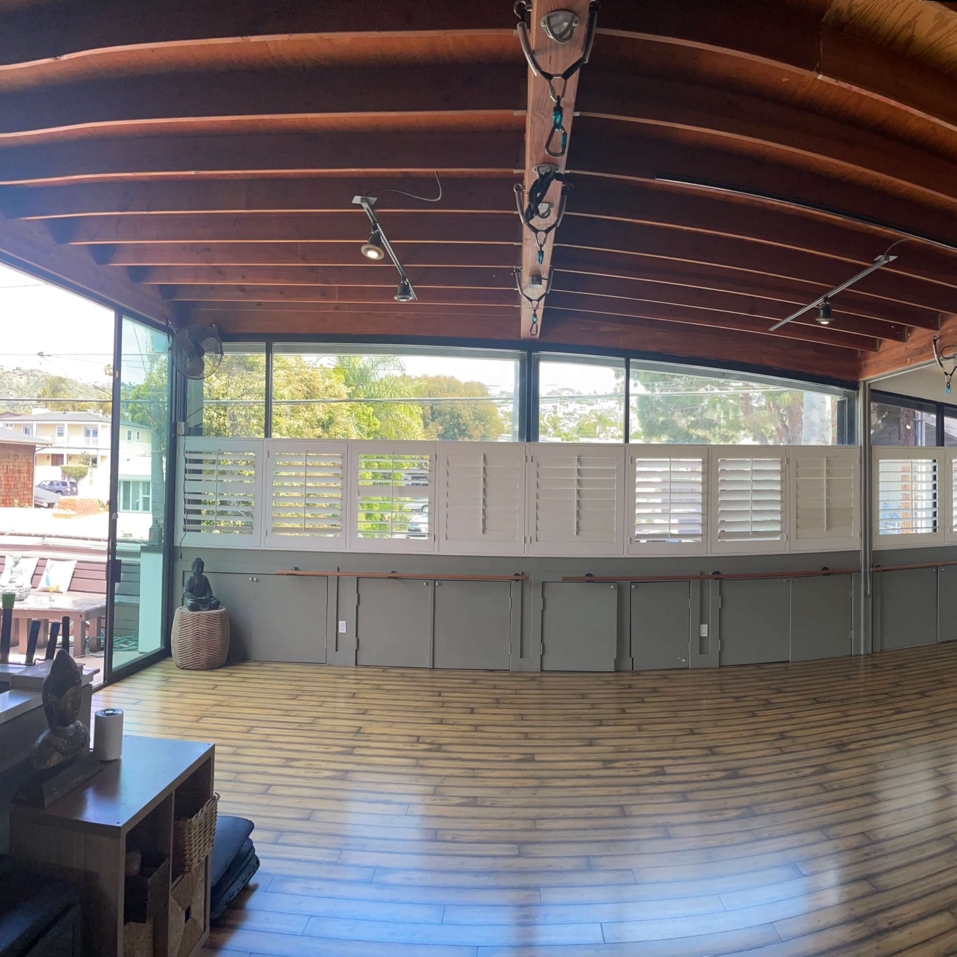 A spacious indoor area with wooden beams on the ceiling, large windows, and a wooden floor, featuring partitioned walls with shutters.