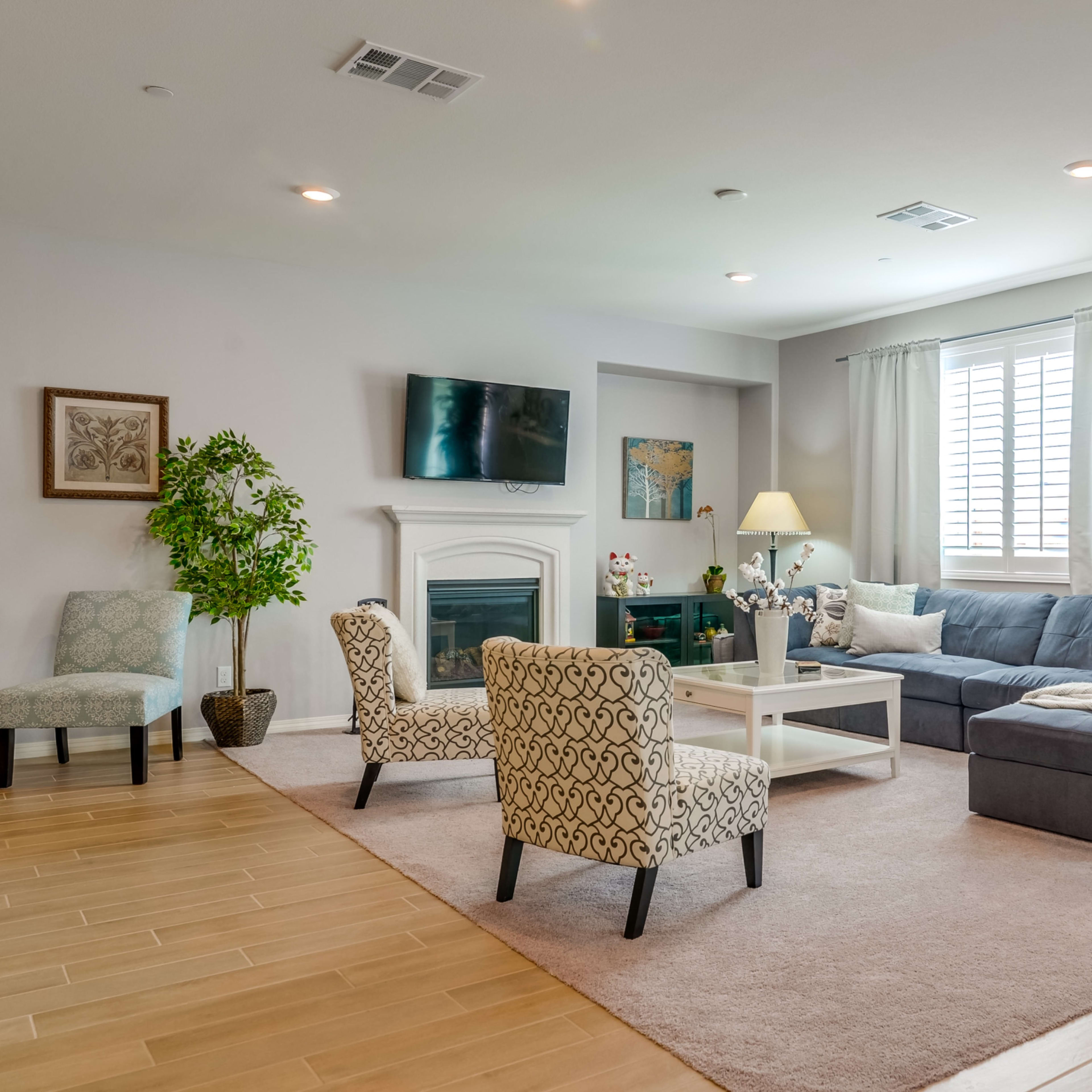 A spacious living room with a blue sectional sofa, a coffee table, and two patterned chairs, along with a fireplace and a large window.