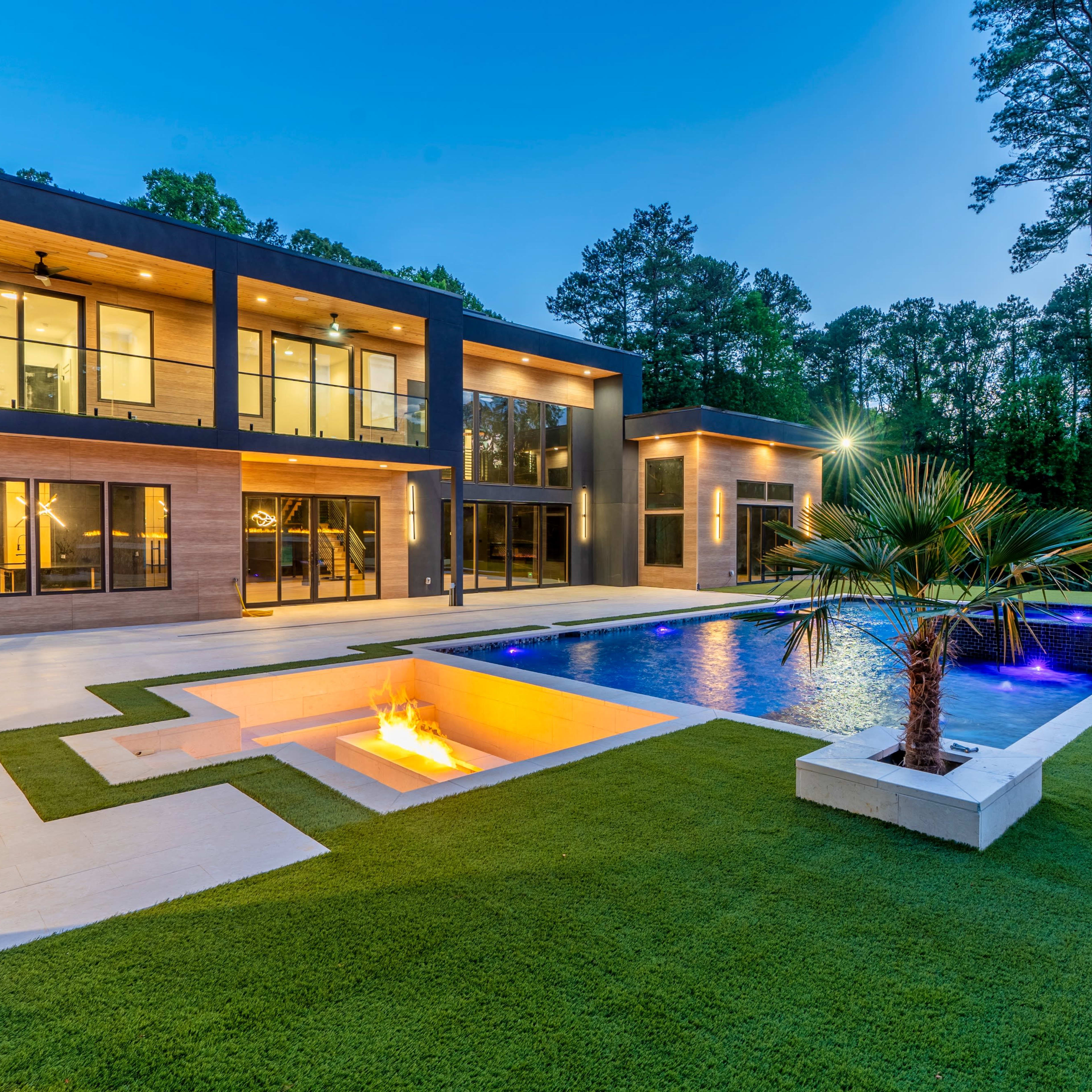 A modern house with large windows overlooks a pool surrounded by grass and palm trees, illuminated by lights in the evening.