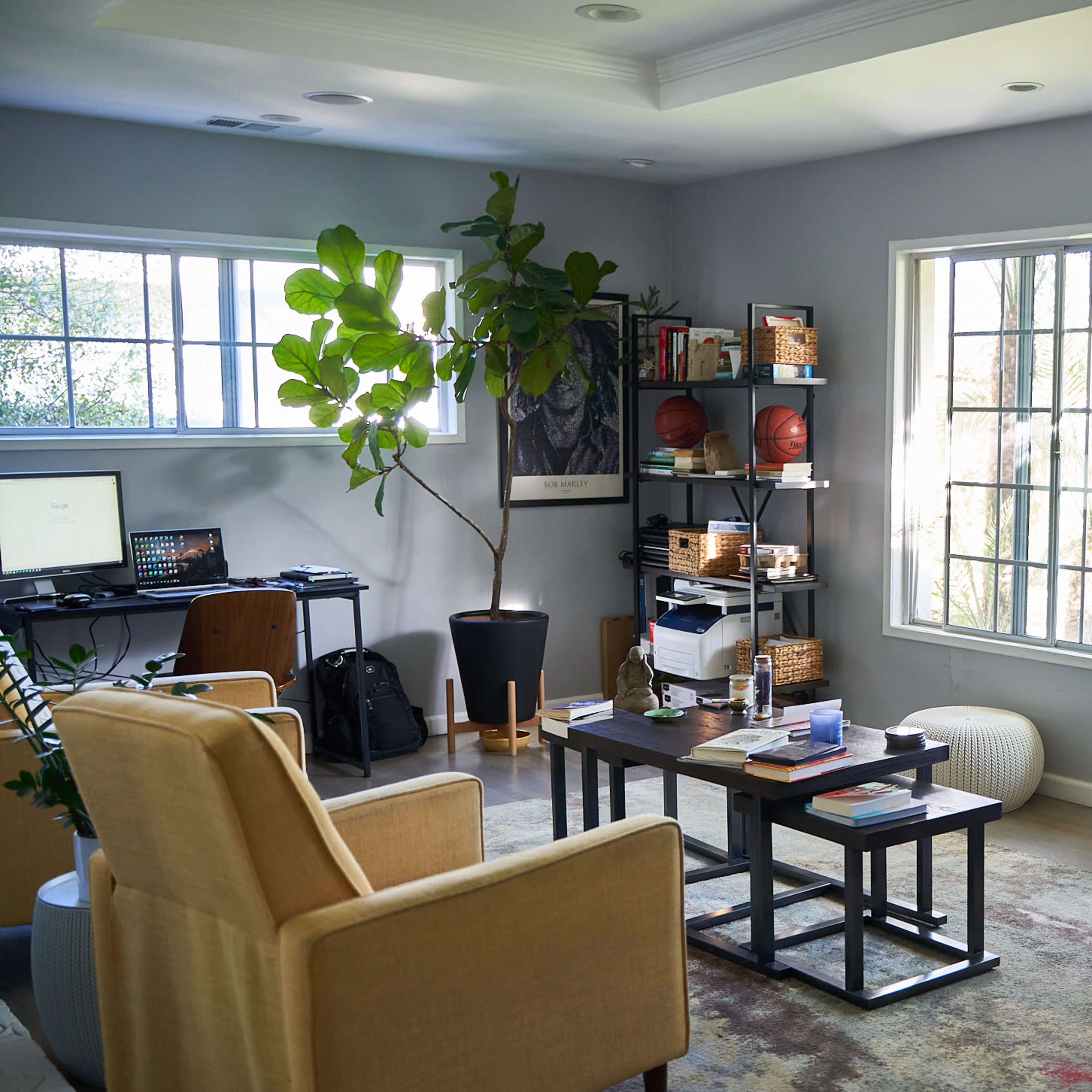 A modern living space features a desk with a computer, shelves filled with books and sports equipment, and large windows letting in natural light.