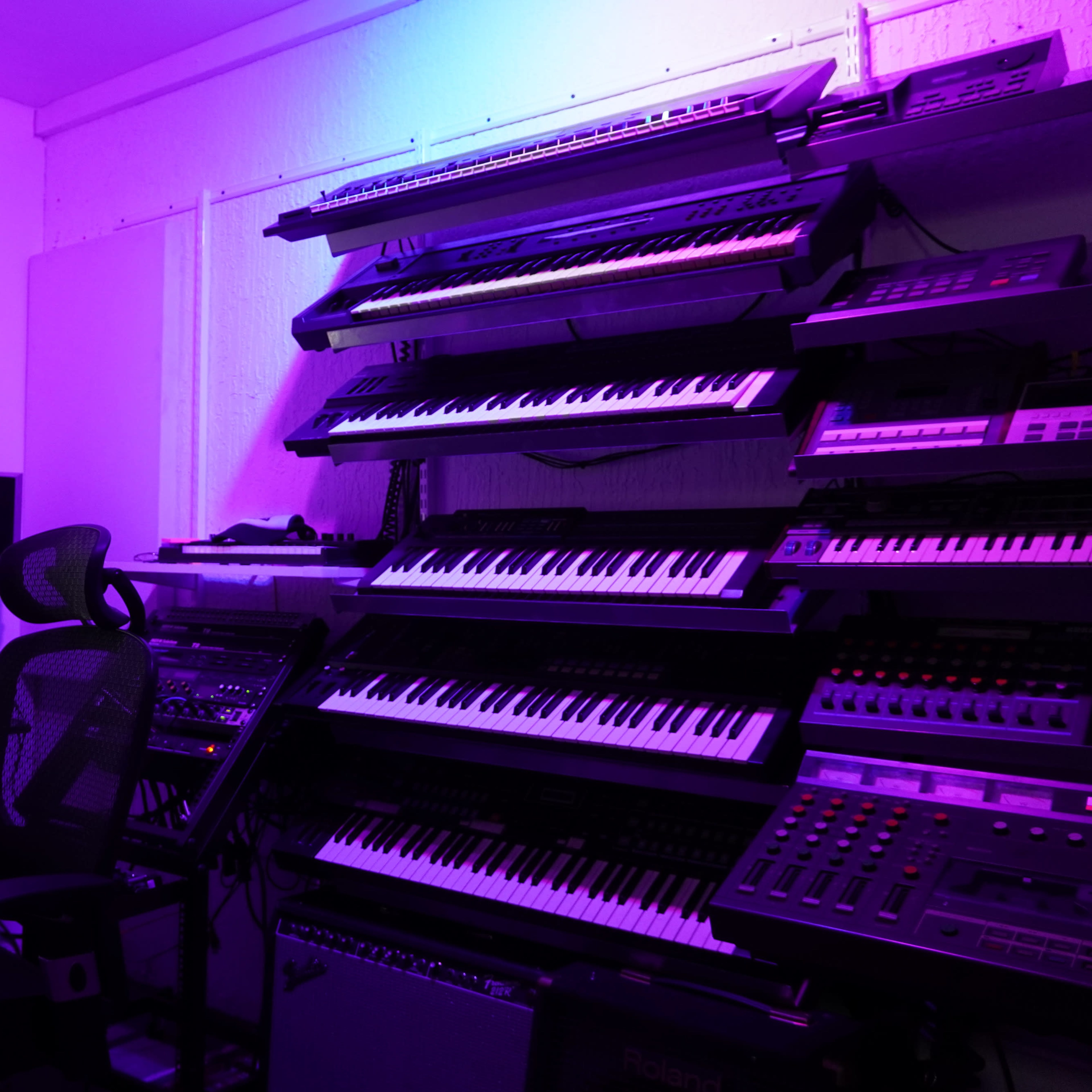 A music production studio features multiple keyboards arranged on shelves with a computer and audio equipment.