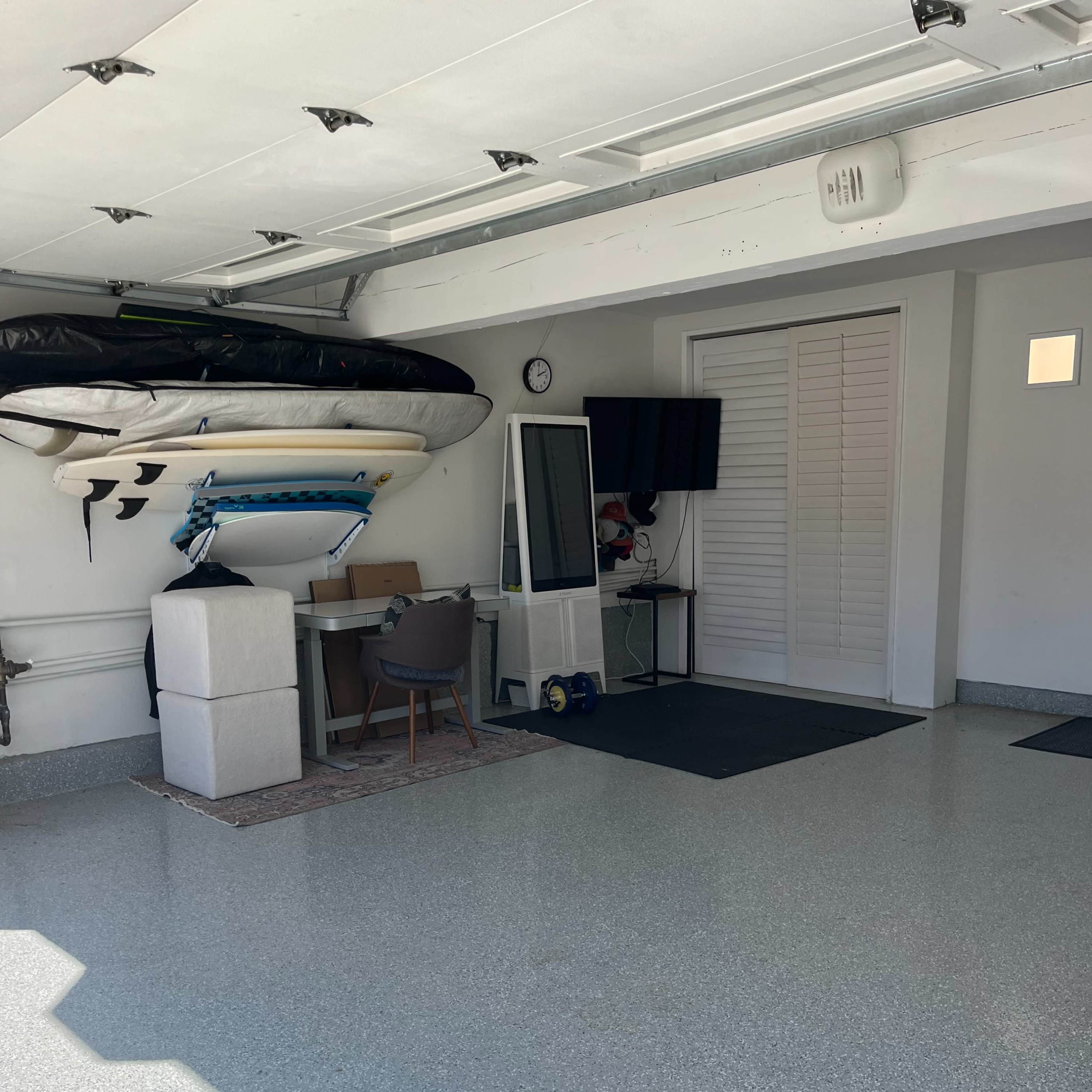 A garage features a mounted surfboard, a desk with a chair, and a television, with a polished floor and light-colored walls.