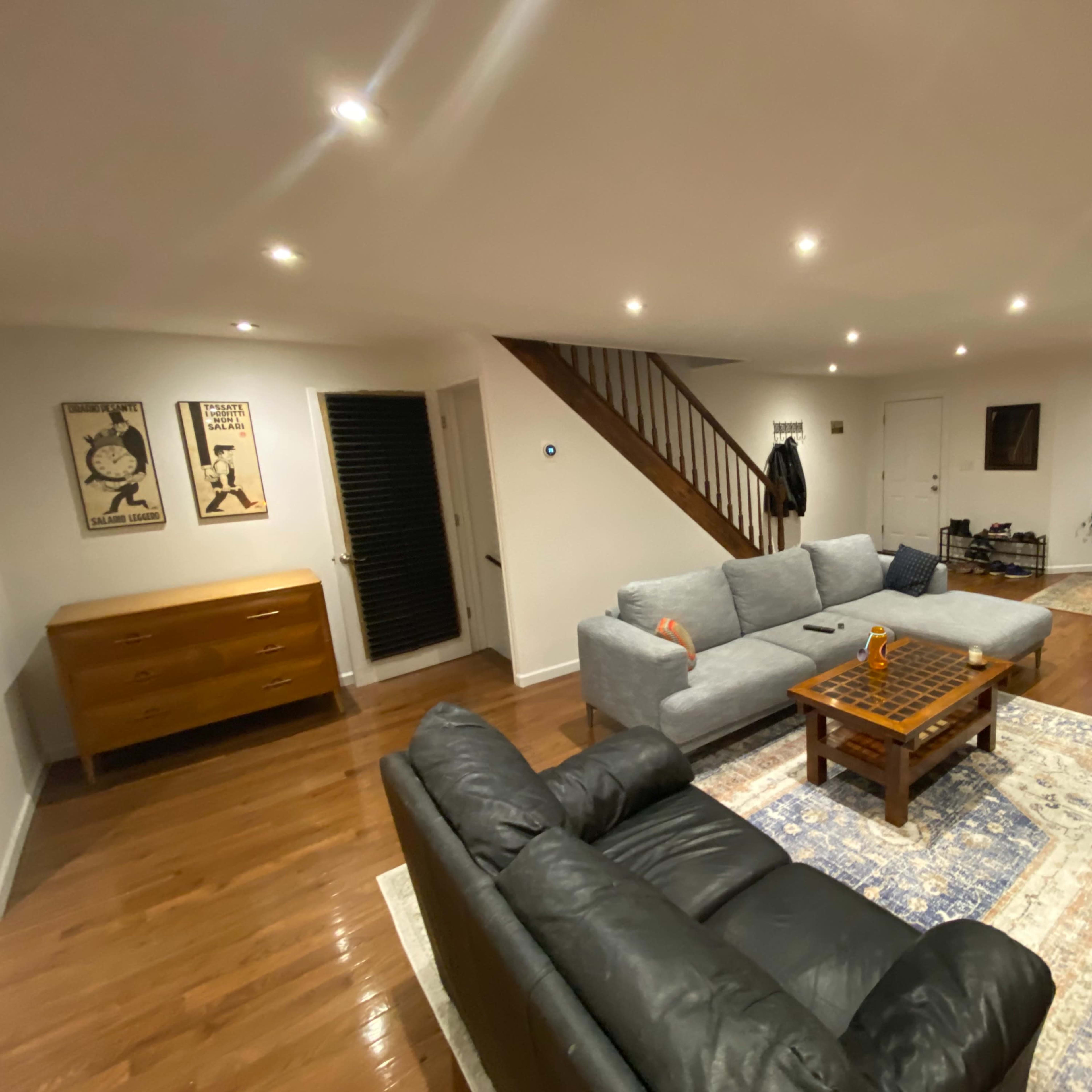 A spacious living room with two couches facing a wooden coffee table, illuminated by recessed lighting, and featuring a staircase in the background.