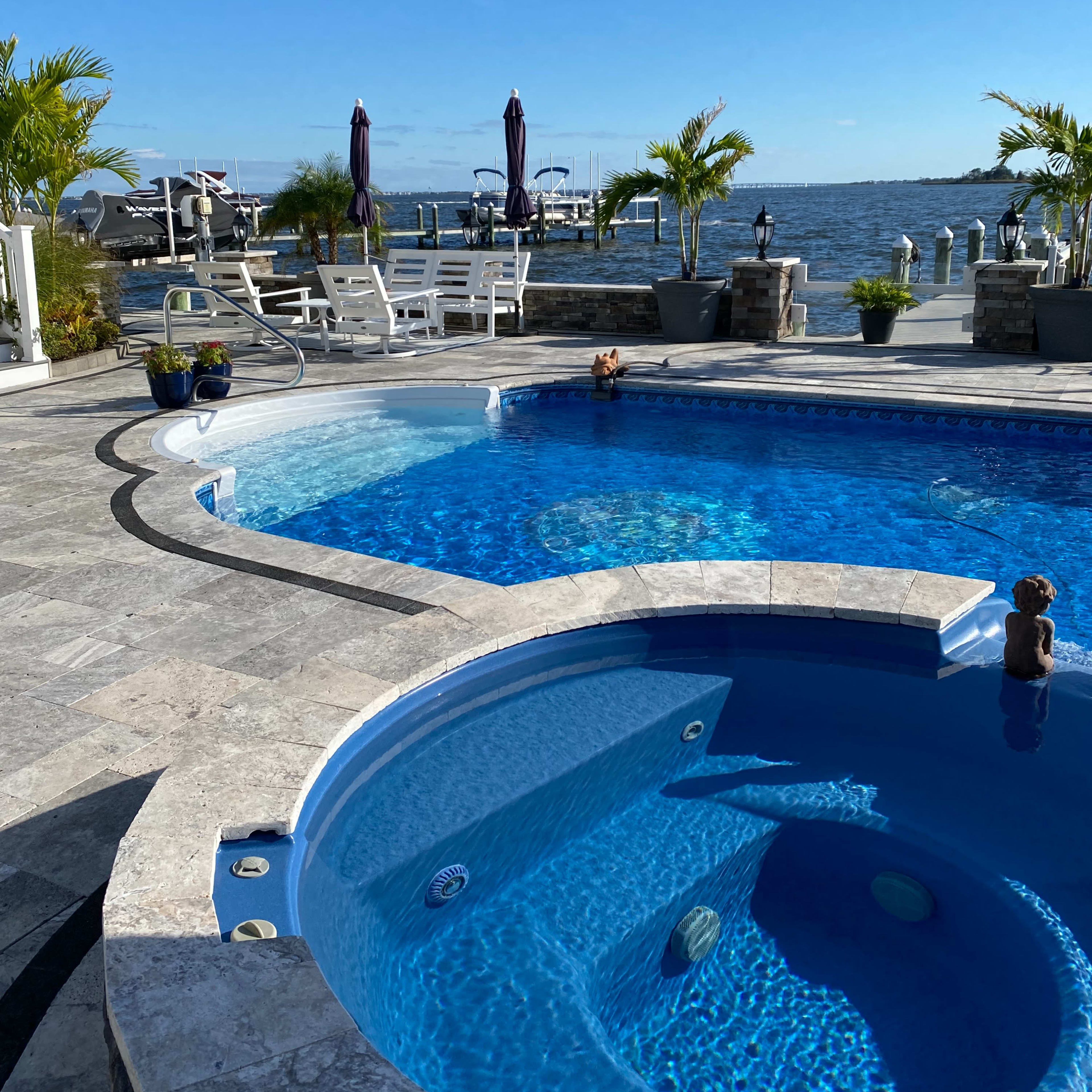 A swimming pool area with a spa, surrounded by stone paving and overlooking a body of water with boats in the background.