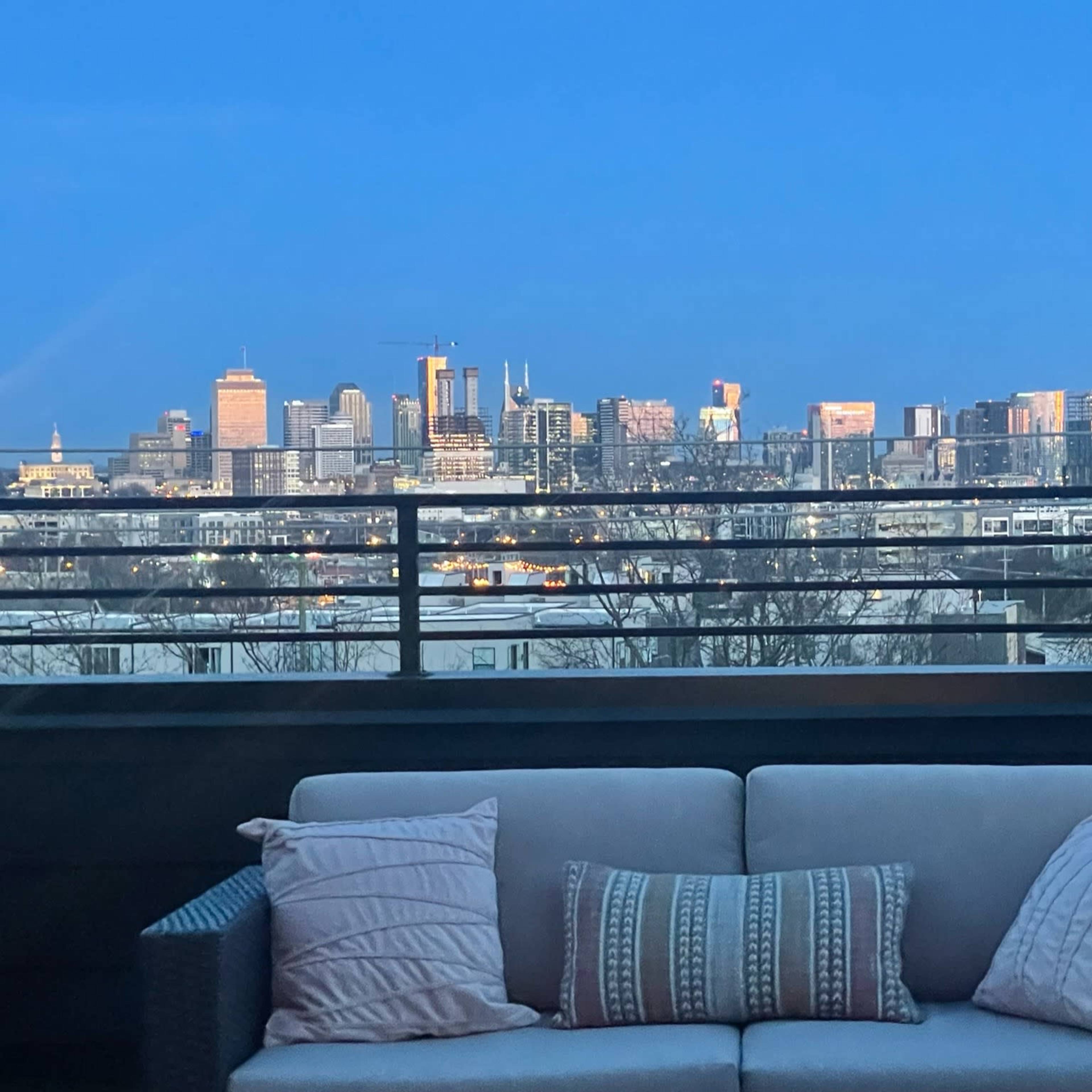 A balcony features a sofa facing a city skyline illuminated at dusk.