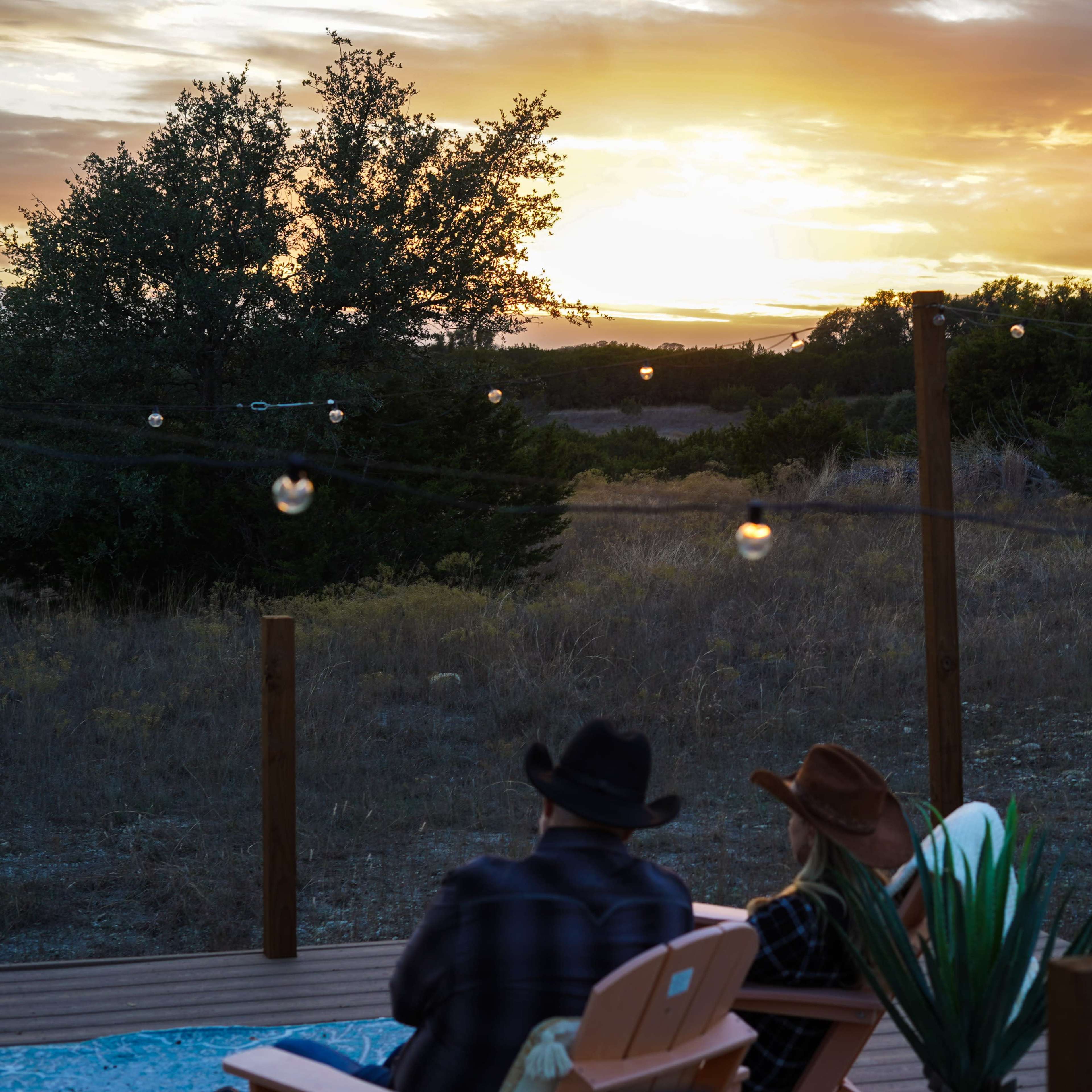 Two people in cowboy hats sit on a deck overlooking a sunset with string lights hanging nearby.