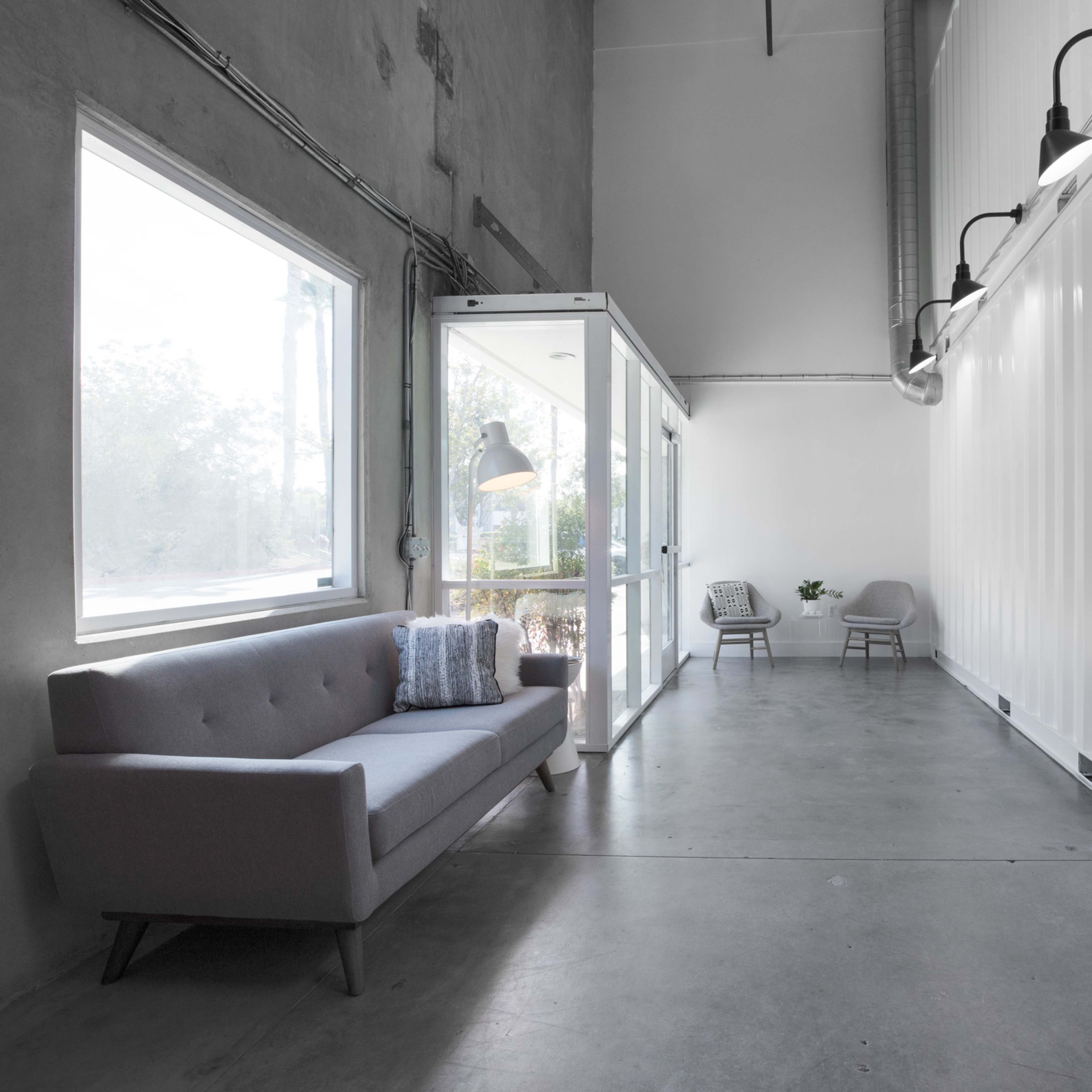 A modern interior space featuring a gray sofa, two chairs, and large windows, with a corrugated shipping container on one side.