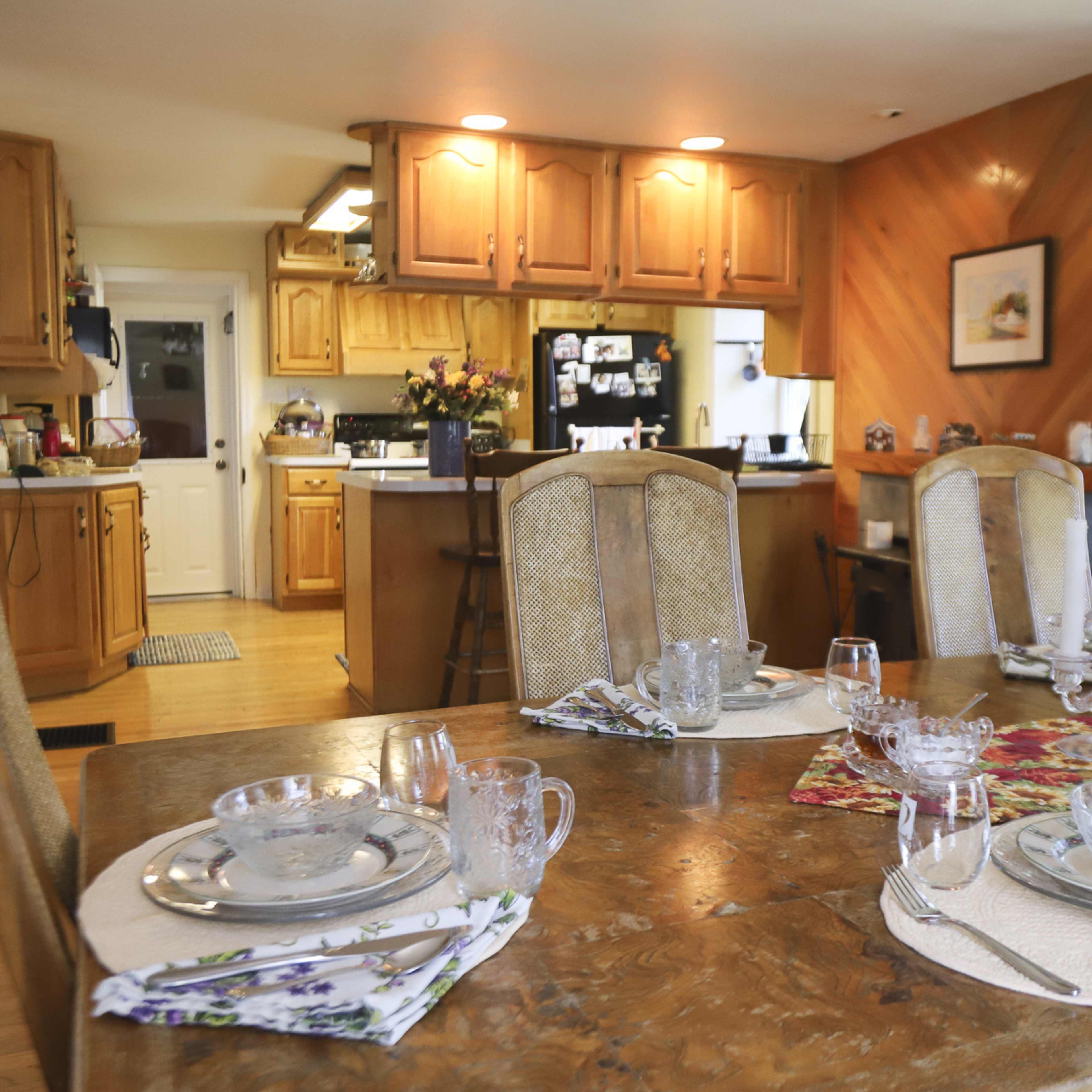A dining table is set with glassware and place settings, with an open kitchen visible in the background.