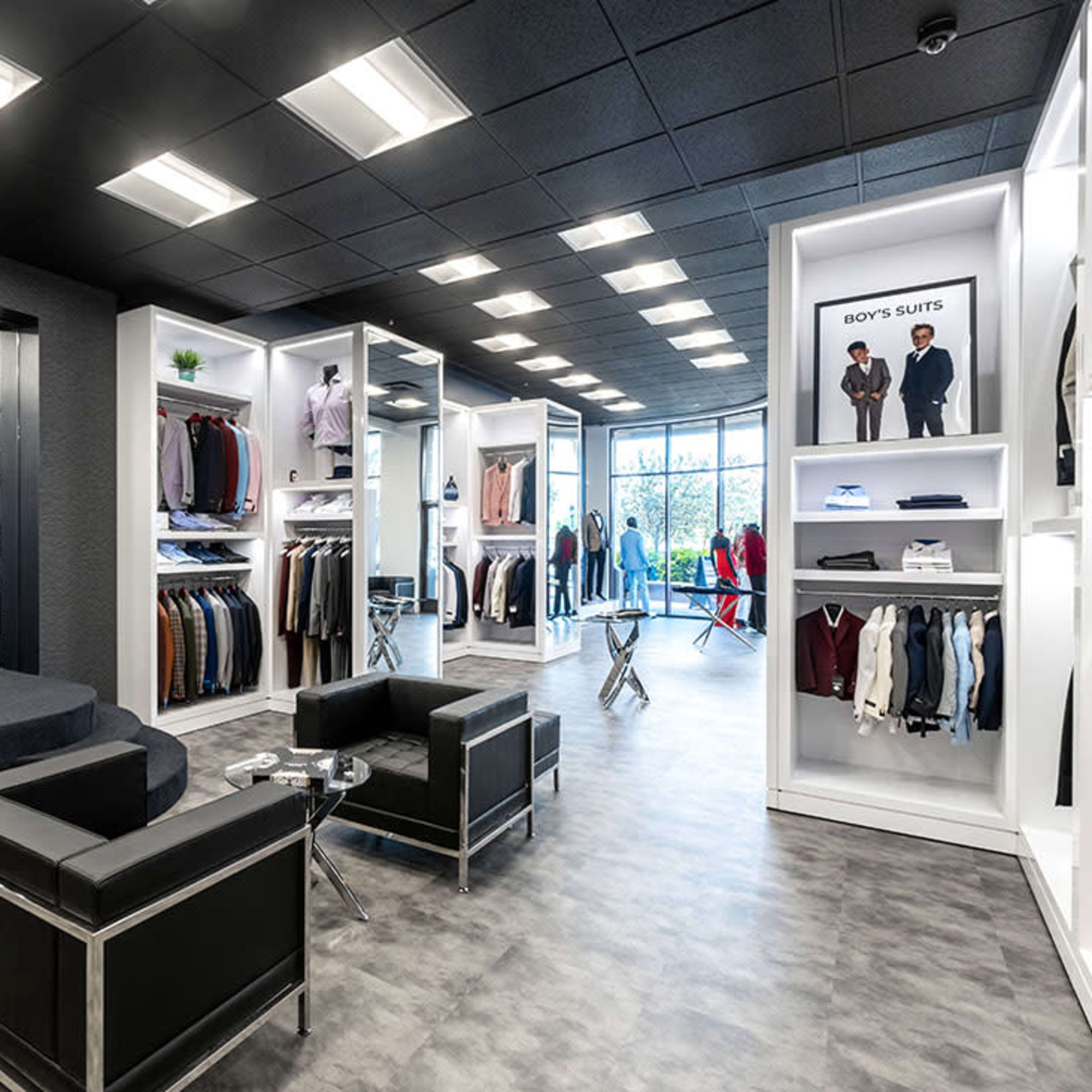 A modern retail clothing store featuring white display units with various garments, a seating area with black chairs, and large windows allowing natural light.