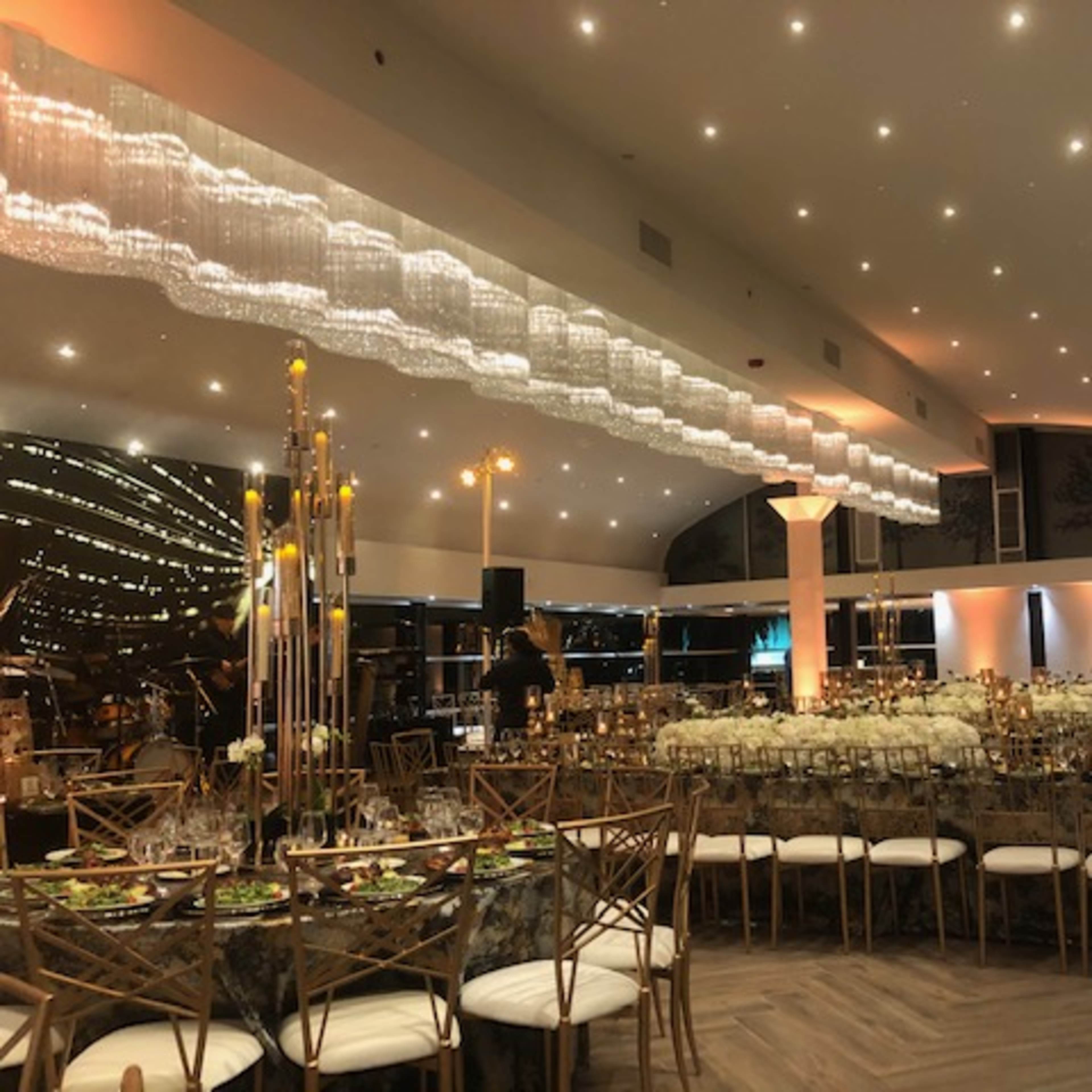 An outdoor venue at night, featuring a wooden floor, illuminated trees, string lights overhead, and modern glass structures.
