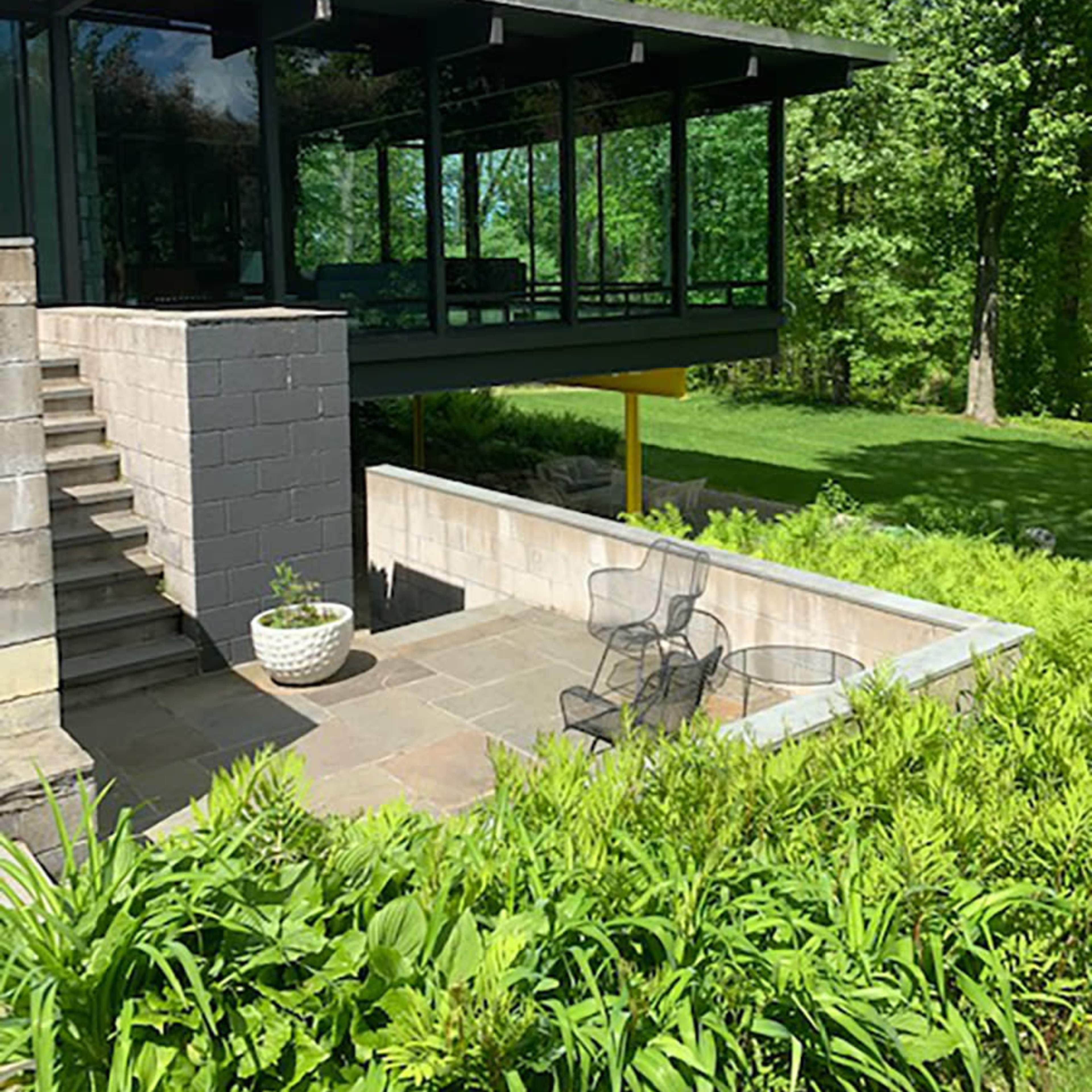 A modern glass house is elevated on a concrete base, surrounded by greenery and featuring steps leading to a landscaped area.