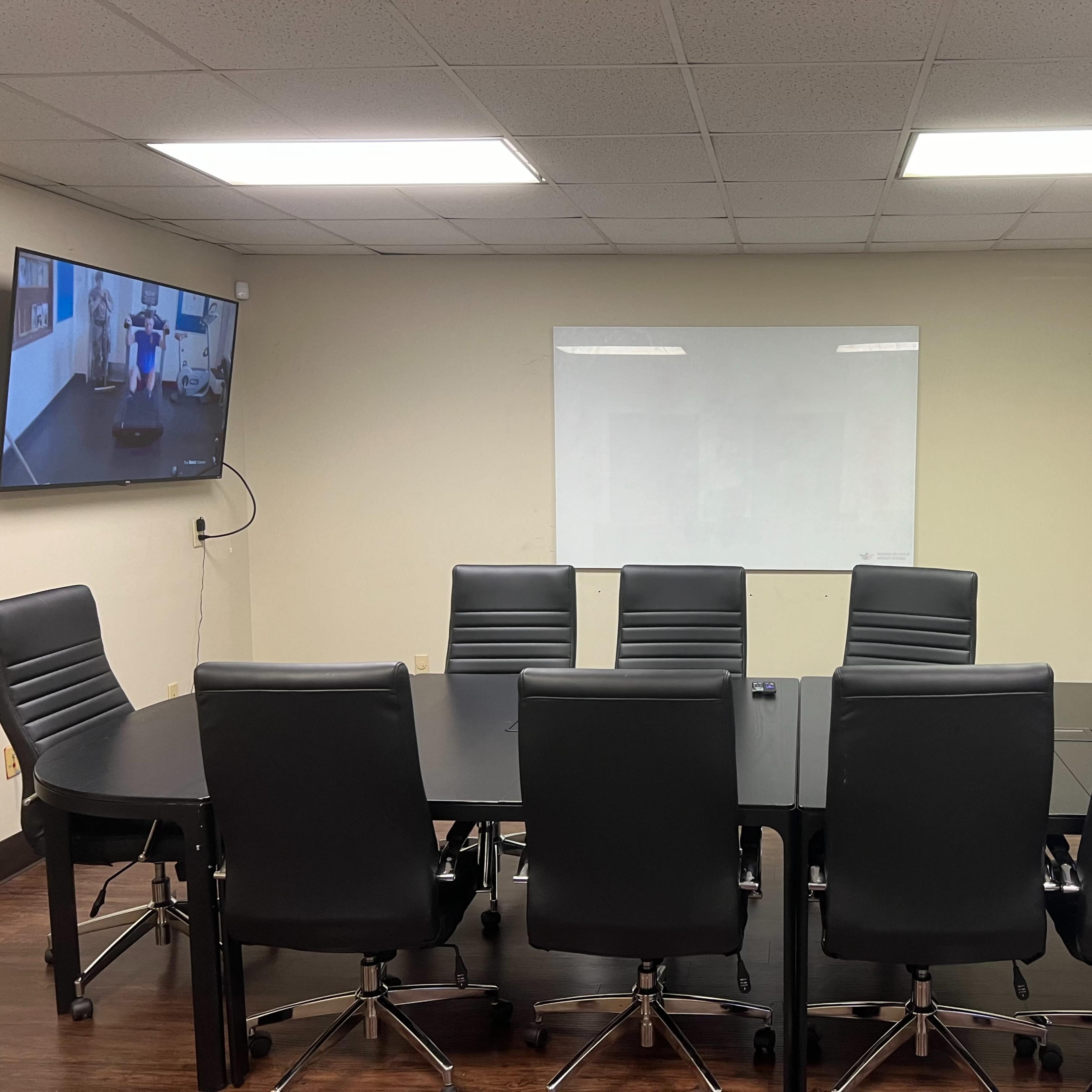 A conference room with a large black table surrounded by eight black leather chairs and a wall-mounted screen displaying a video.