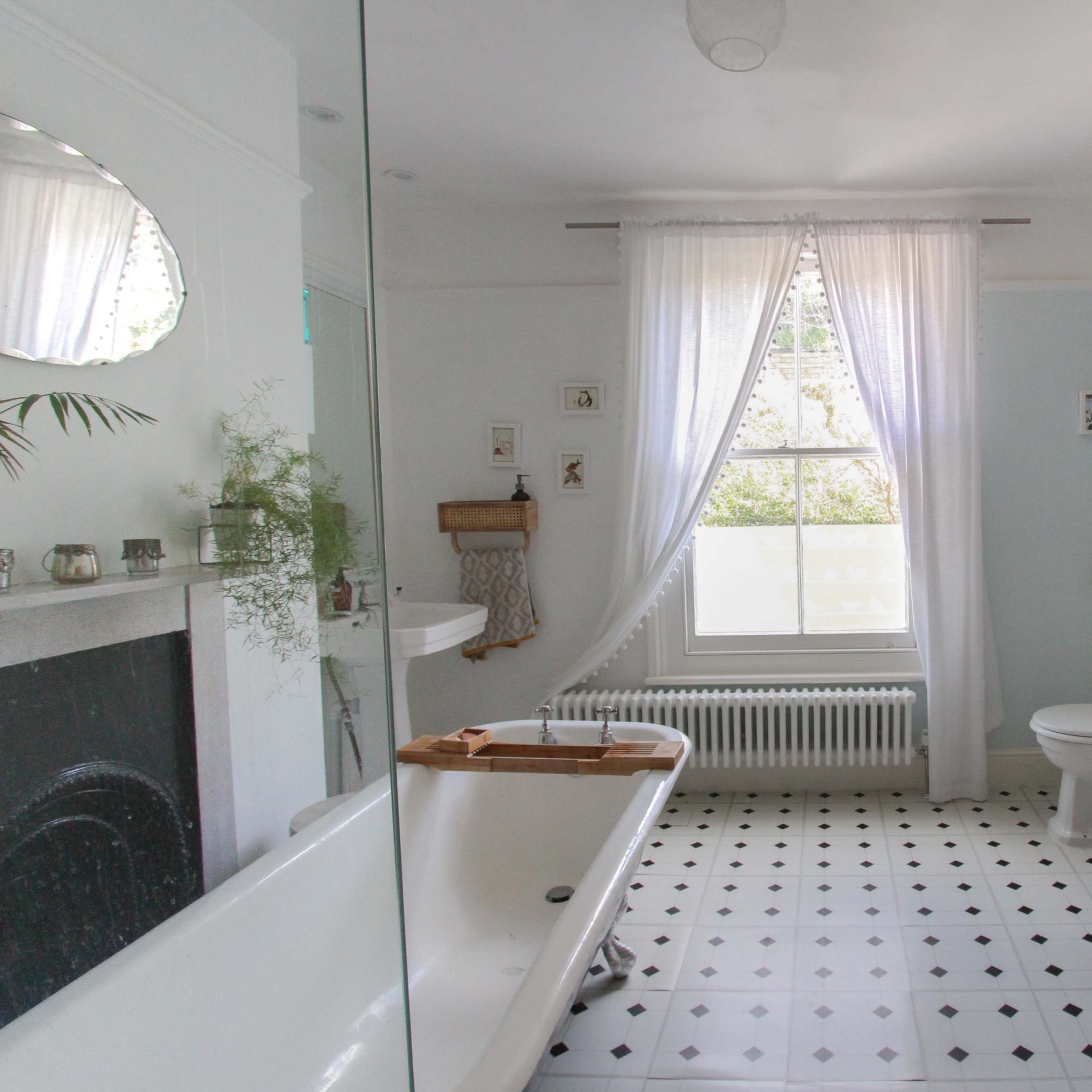 A light-colored bathroom featuring a freestanding bathtub, a window with white curtains, a toilet, and a decorative chair on a tiled floor.