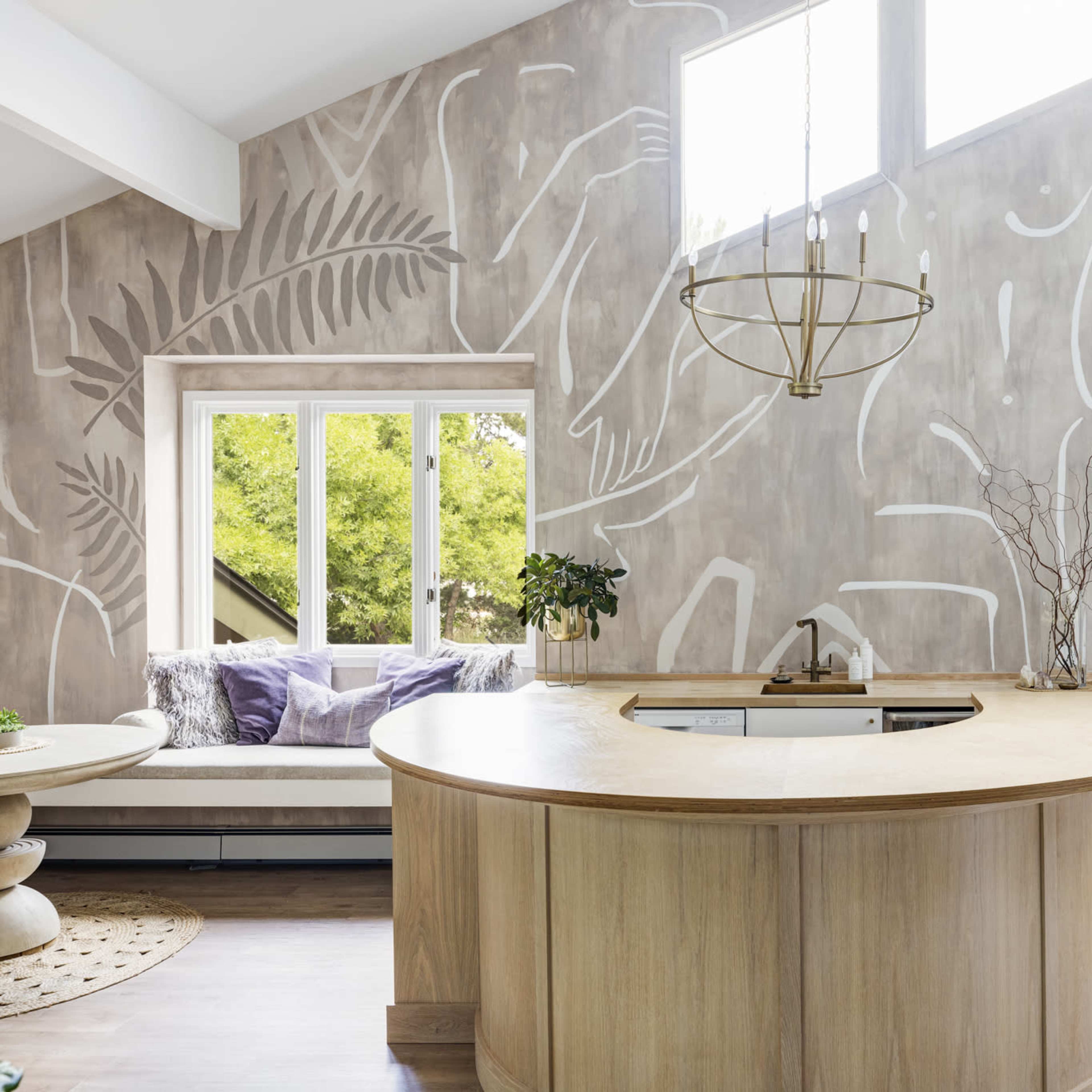 A modern kitchen with a curved wooden countertop, surrounded by a wall adorned with abstract white line art and a large window providing natural light.