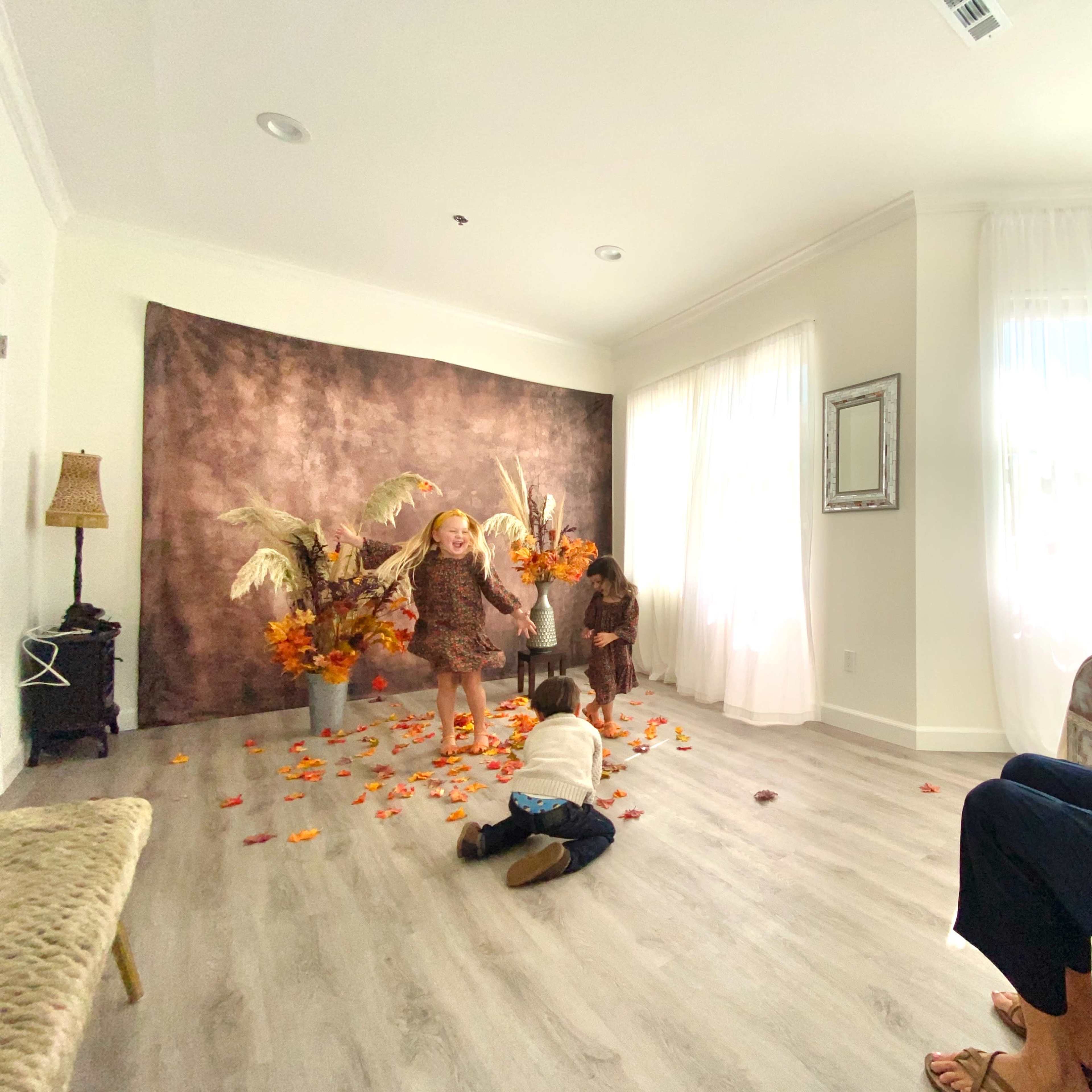 A photographer captures children playing in front of a backdrop adorned with autumn decorations, while leaves are scattered on the floor.